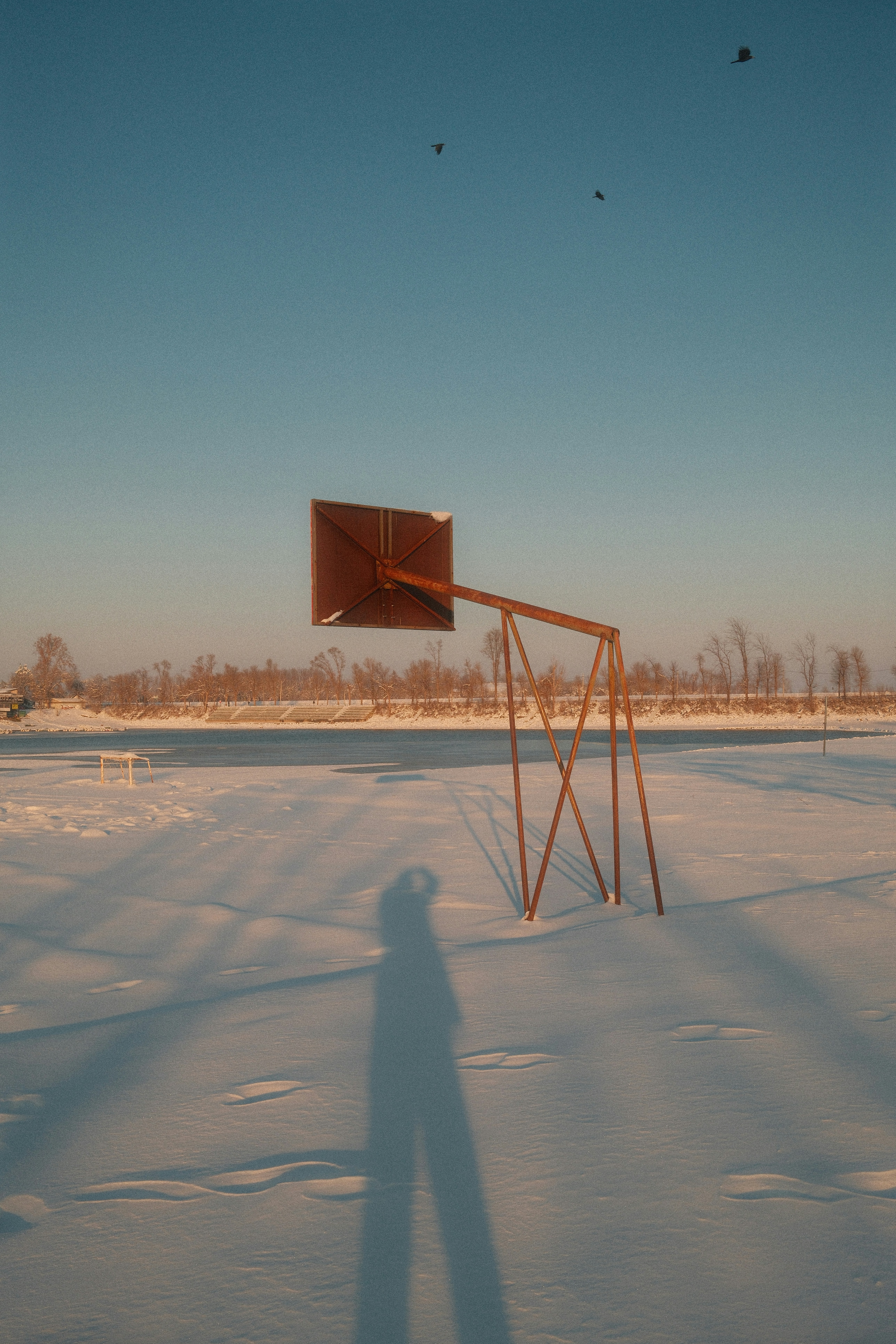 Rusty basketball hoop stands in snowy field at sunset.