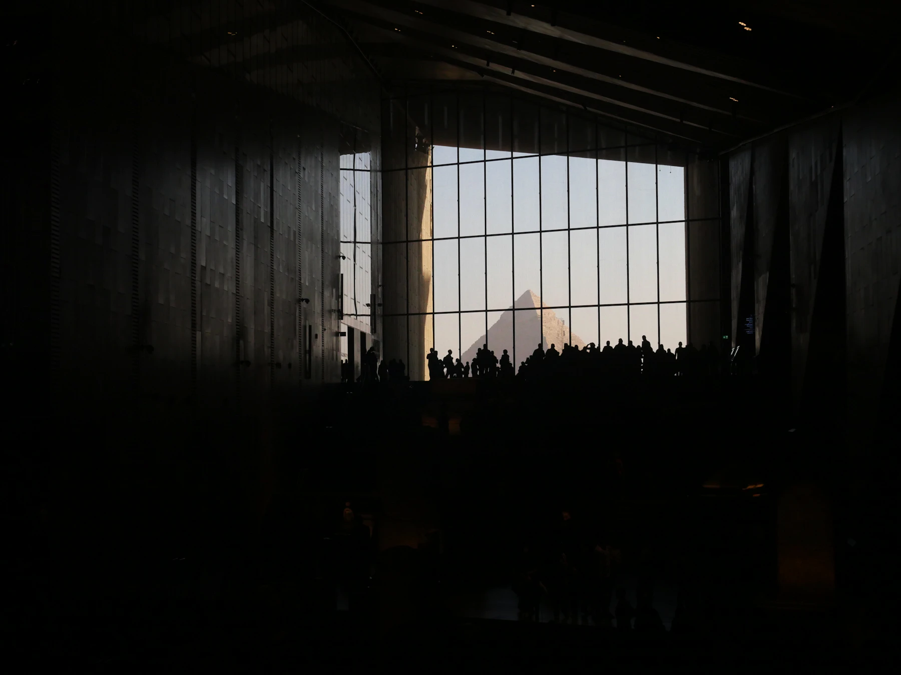 Visitor silhouettes stand before the Grand Egyptian Museum's glazed pyramid view.