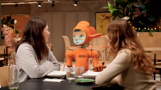 Two women talk to an orange robot at a table.