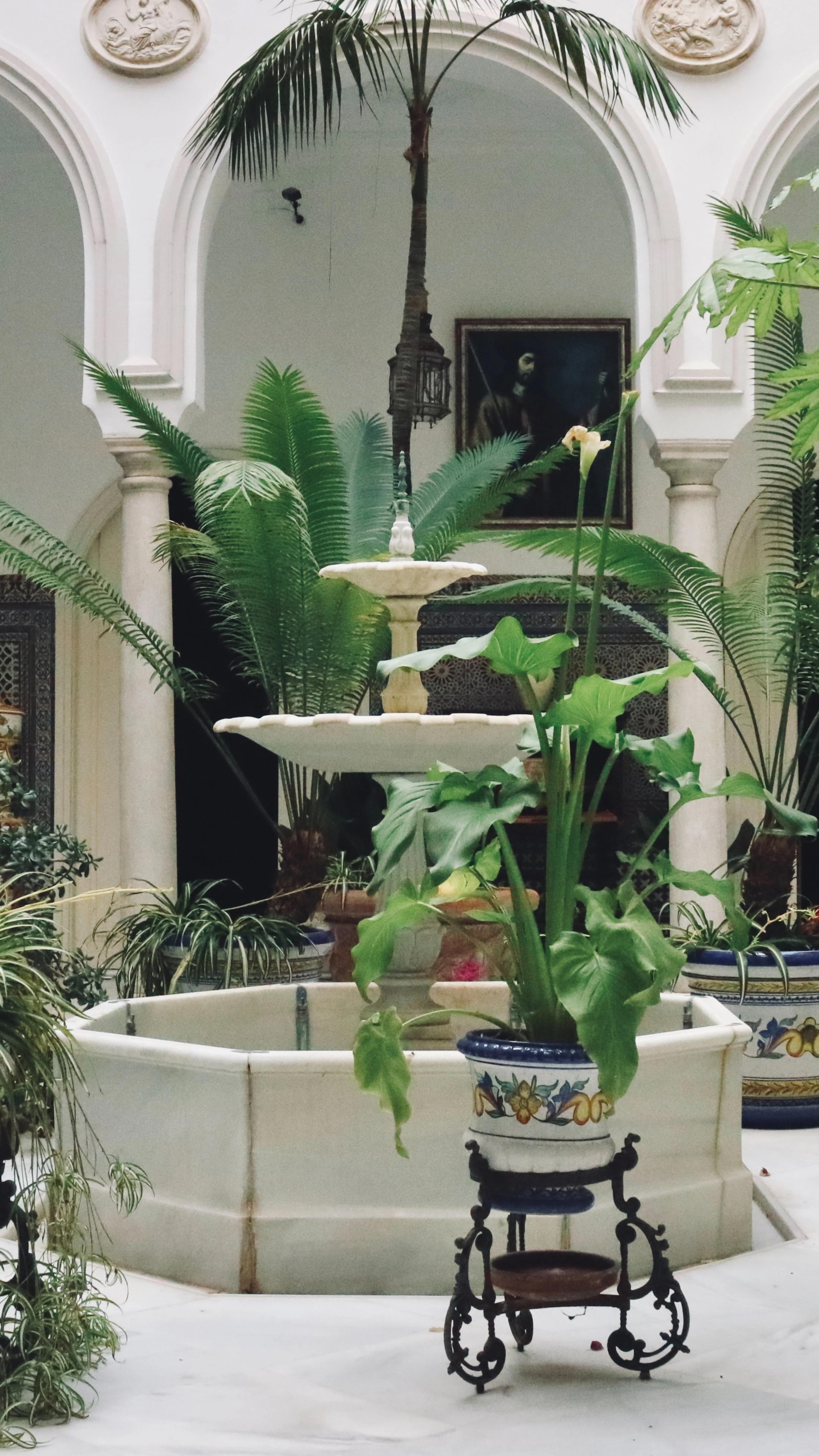 A whitewashed Sevillian courtyard with a tiered marble fountain, palms and ferns in painted ceramic pots, and an old oil portrait hung between the arches.