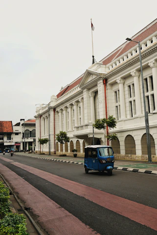 A blue auto rickshaw drives past a white colonial building.