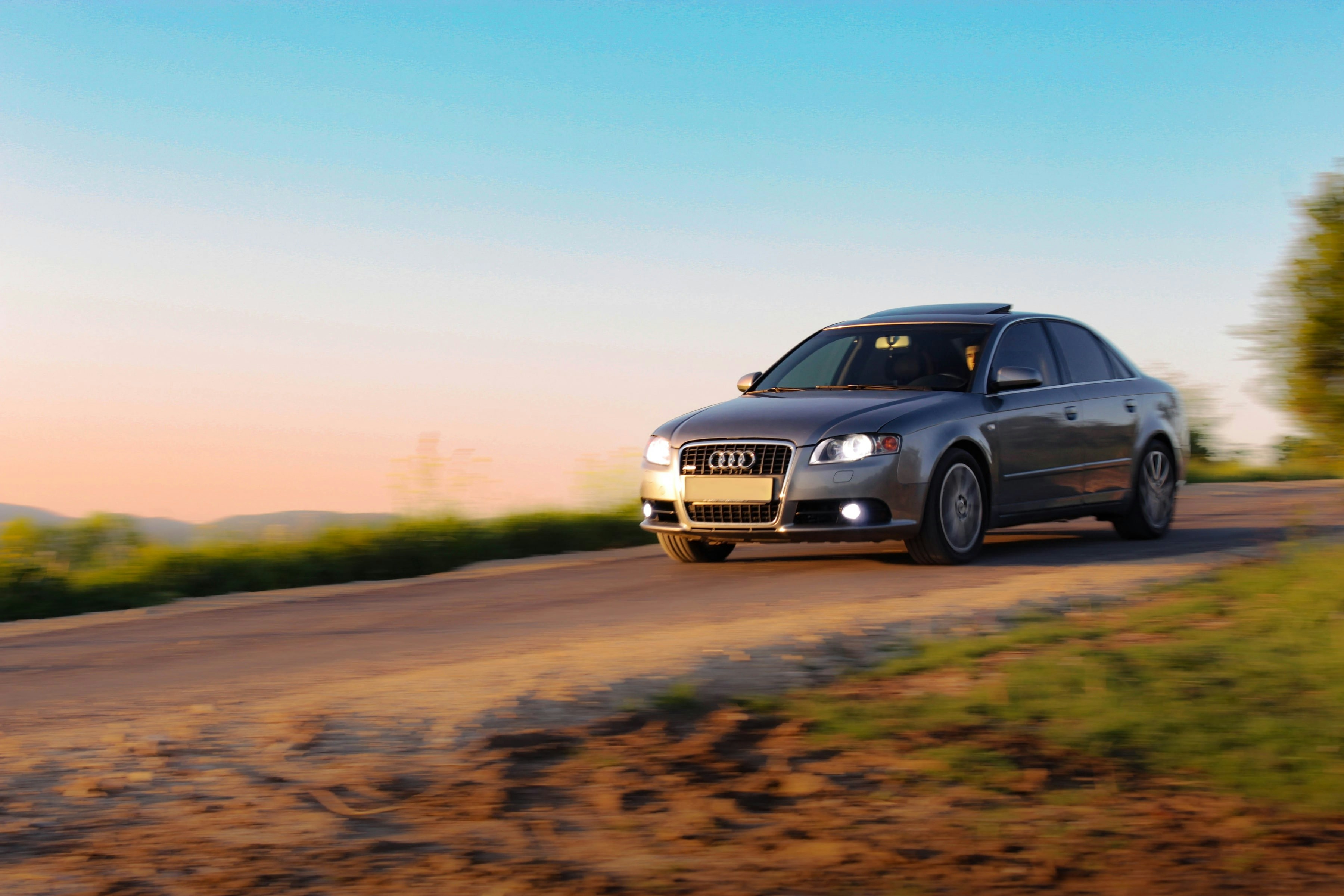 A silver sedan drives on a dirt road at sunset.