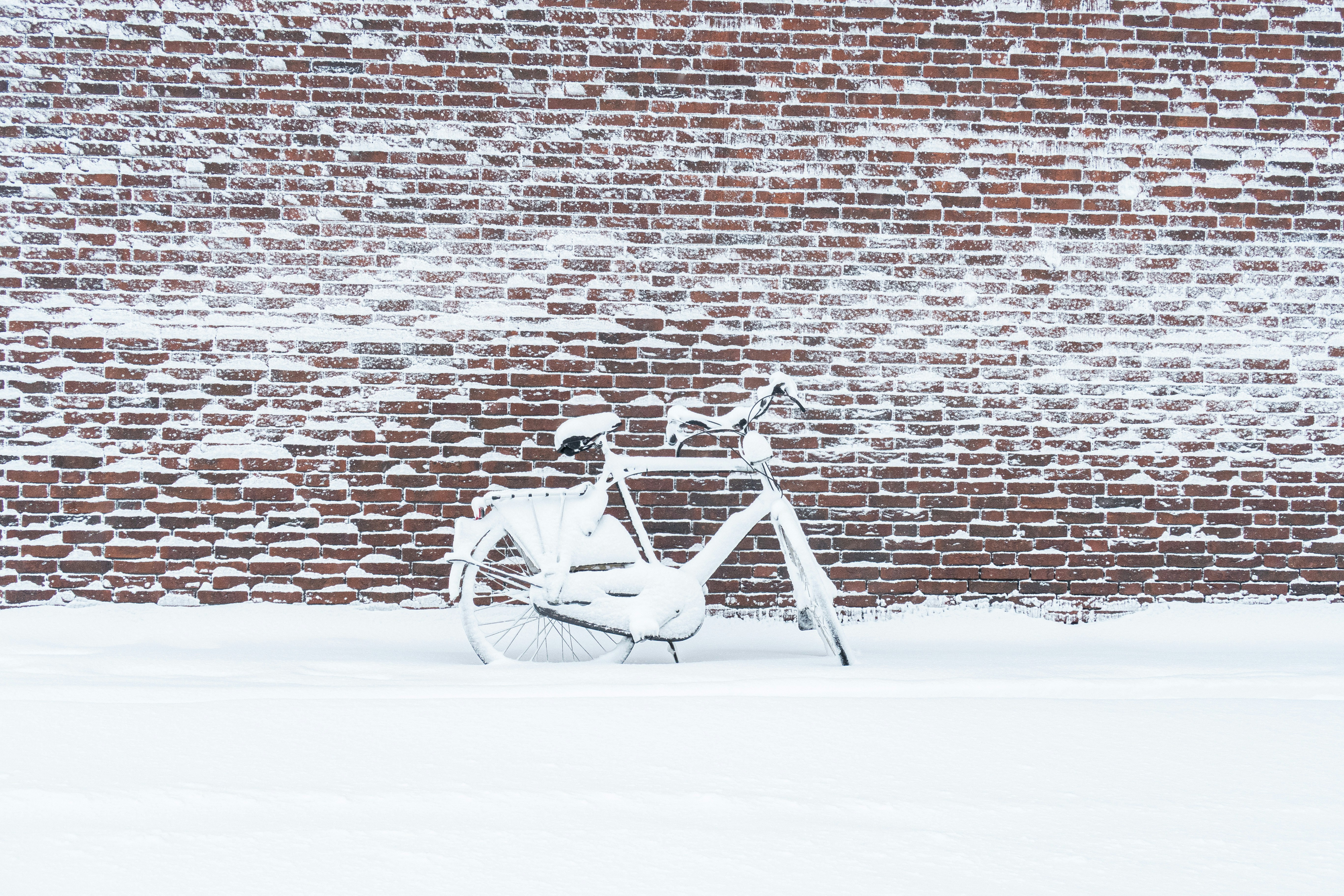 A white bicycle covered in snow against a brick wall.