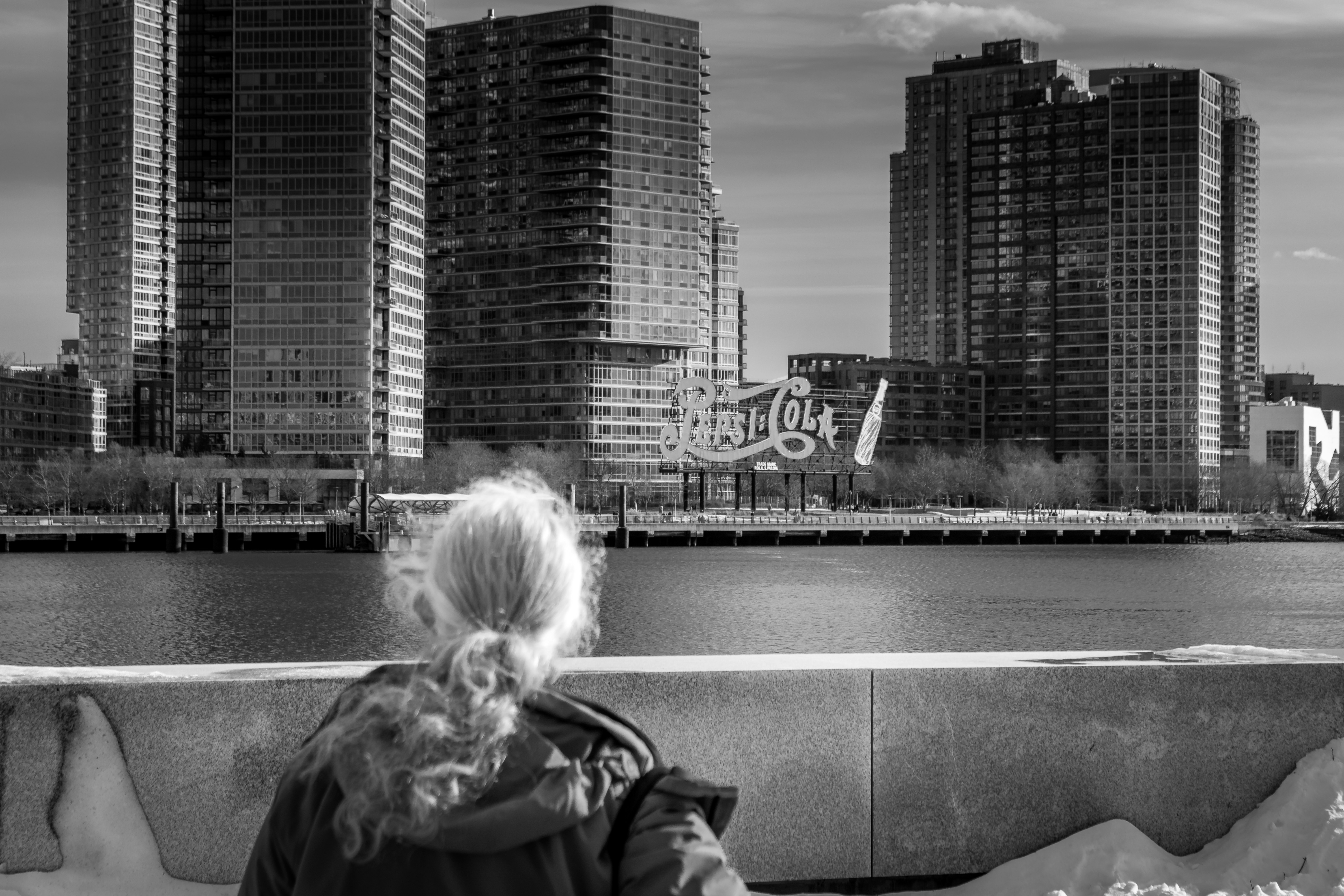 A winter view across the East River shows the Long Island City waterfront in Queens, seen from Roosevelt Island. Modern residential towers line the shoreline, reflecting New York City’s rapid urban development. The iconic Pepsi Cola sign stands along the riverfront, adding historical character against a backdrop of glass high rises. In the foreground, an old lady looks toward the skyline, creating a human scale contrast. Snow, bare trees, and calm water emphasize the seasonal atmosphere and evolving cityscape.