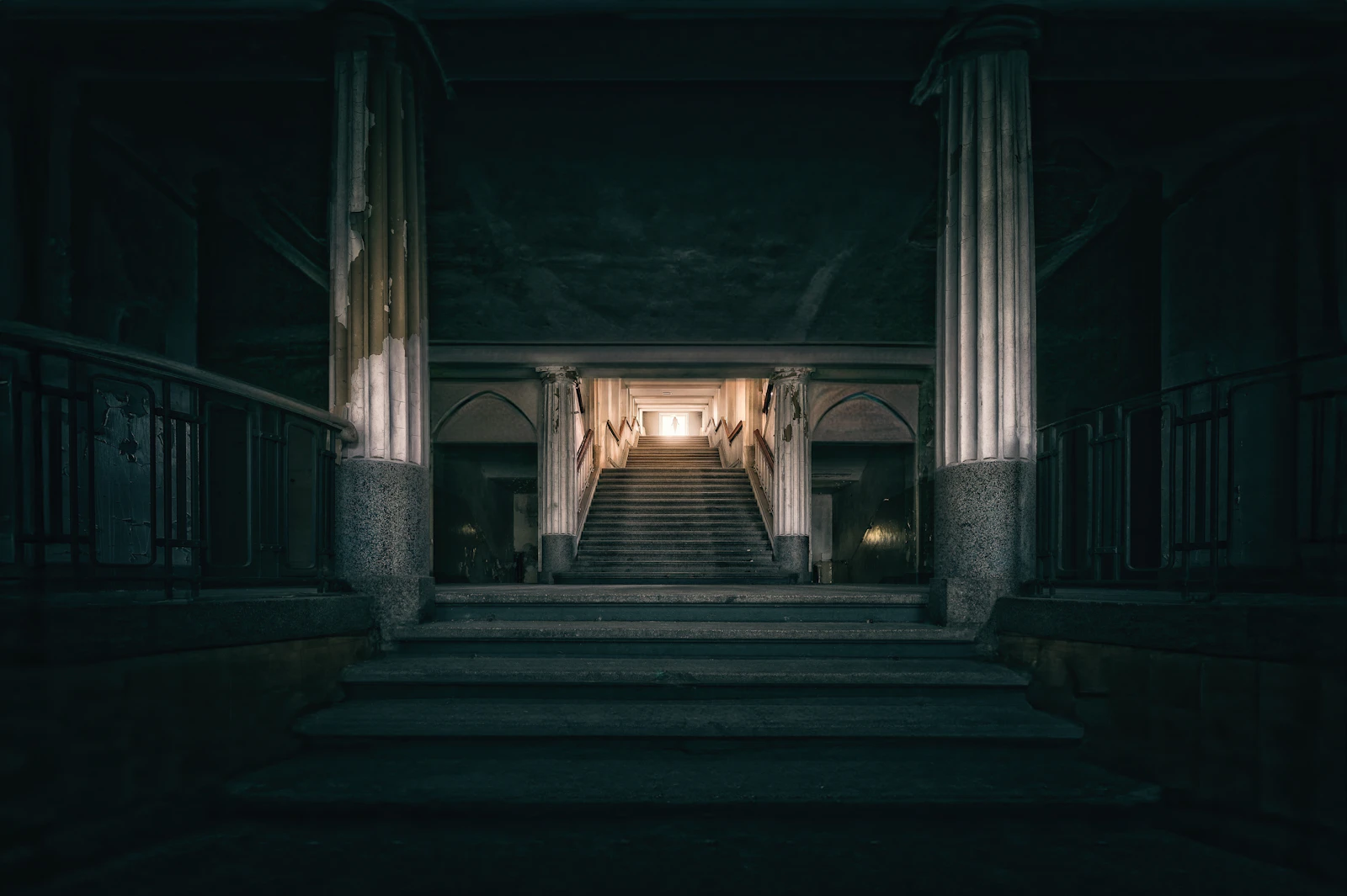 Dim institutional hall with stone columns and a grand staircase rising toward a warmly lit doorway
