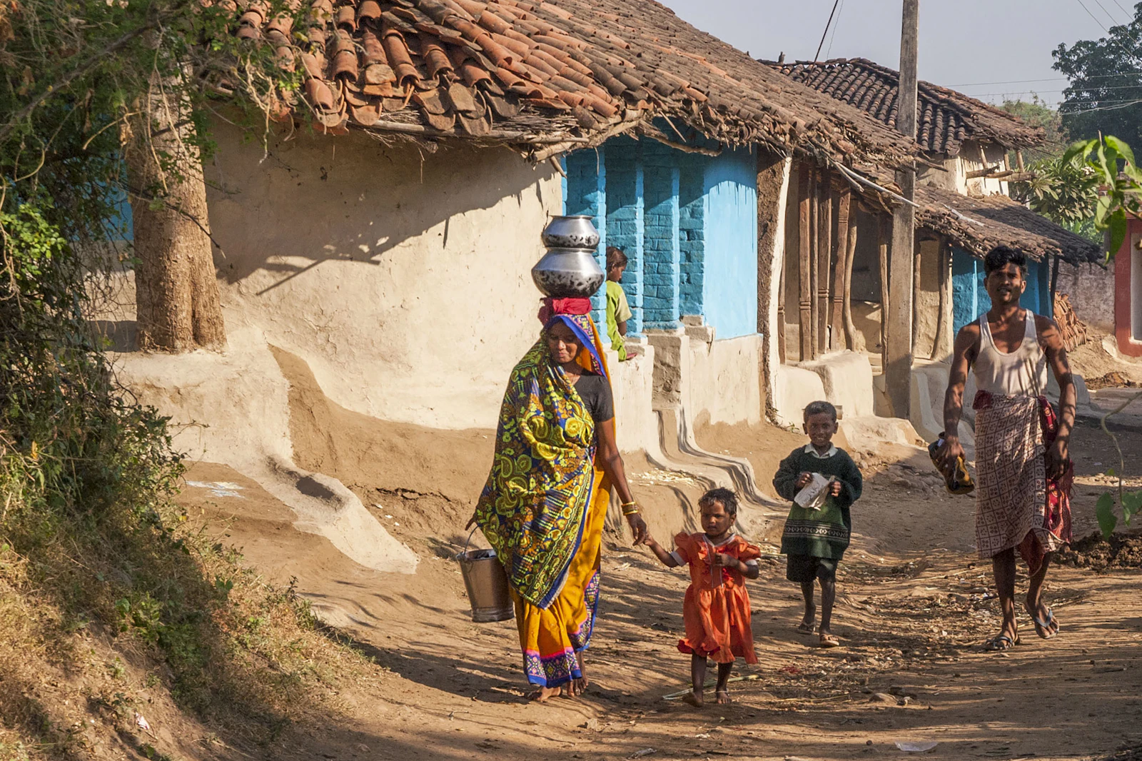 A woman carrying water past houses while children play in a lane
