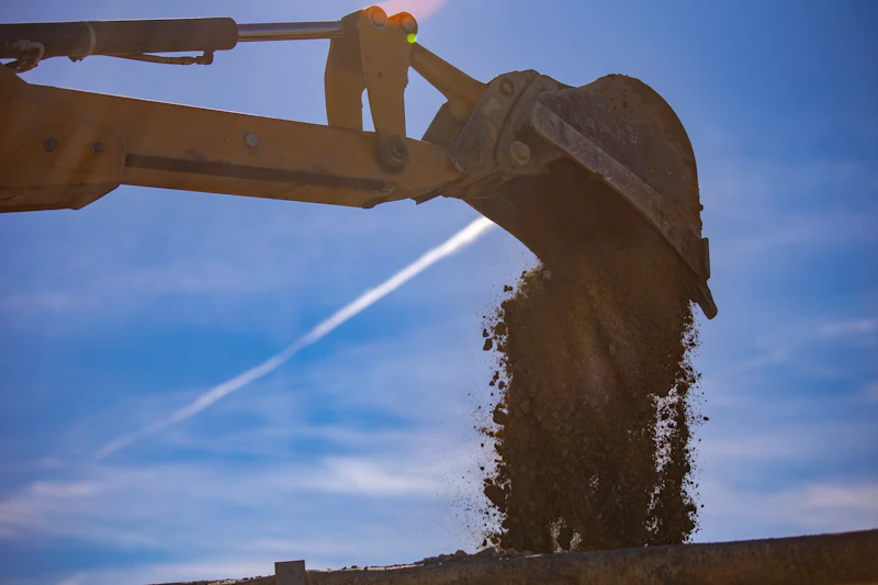 Excavator bucket dumping dirt against a blue sky