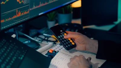 Person using calculator at desk with computer charts.