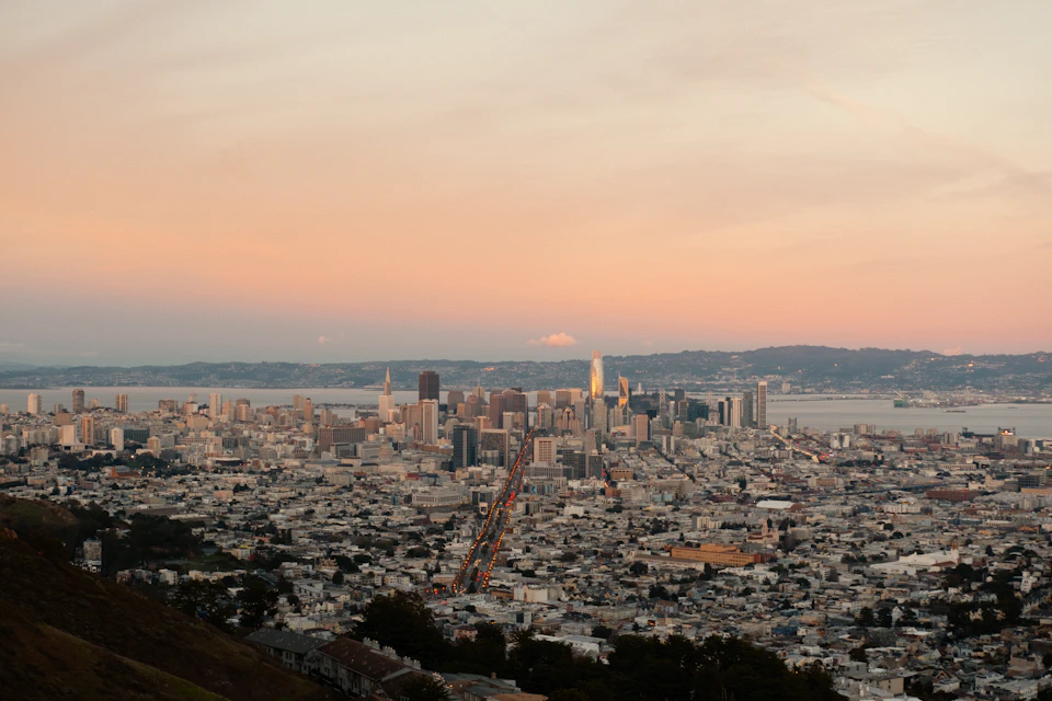 The image presents a panoramic view of the San Francisco Bay Area, with the iconic Golden Gate Bridge spanning across it. The bridge is bathed in warm hues of orange and pink, indicating that the photo was taken during sunset or sunrise. The cityscape below features numerous buildings of varying heights, showcasing the urban landscape of San Francisco.

The sky above mirrors the colors of the bridge, with a few clouds scattered throughout. The perspective of the image is from a high vantage point, providing an expansive view of the bay and the city's skyline.