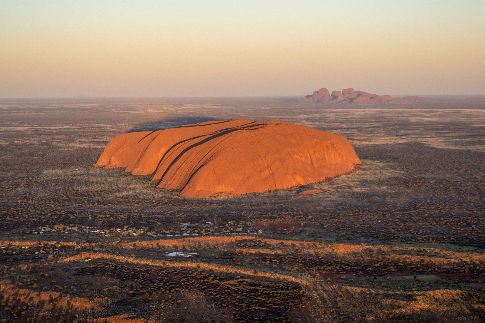 Aerial view of Uluru glowing orange at sunset in the Australian Outback, with Kata Tjuta on the horizon
