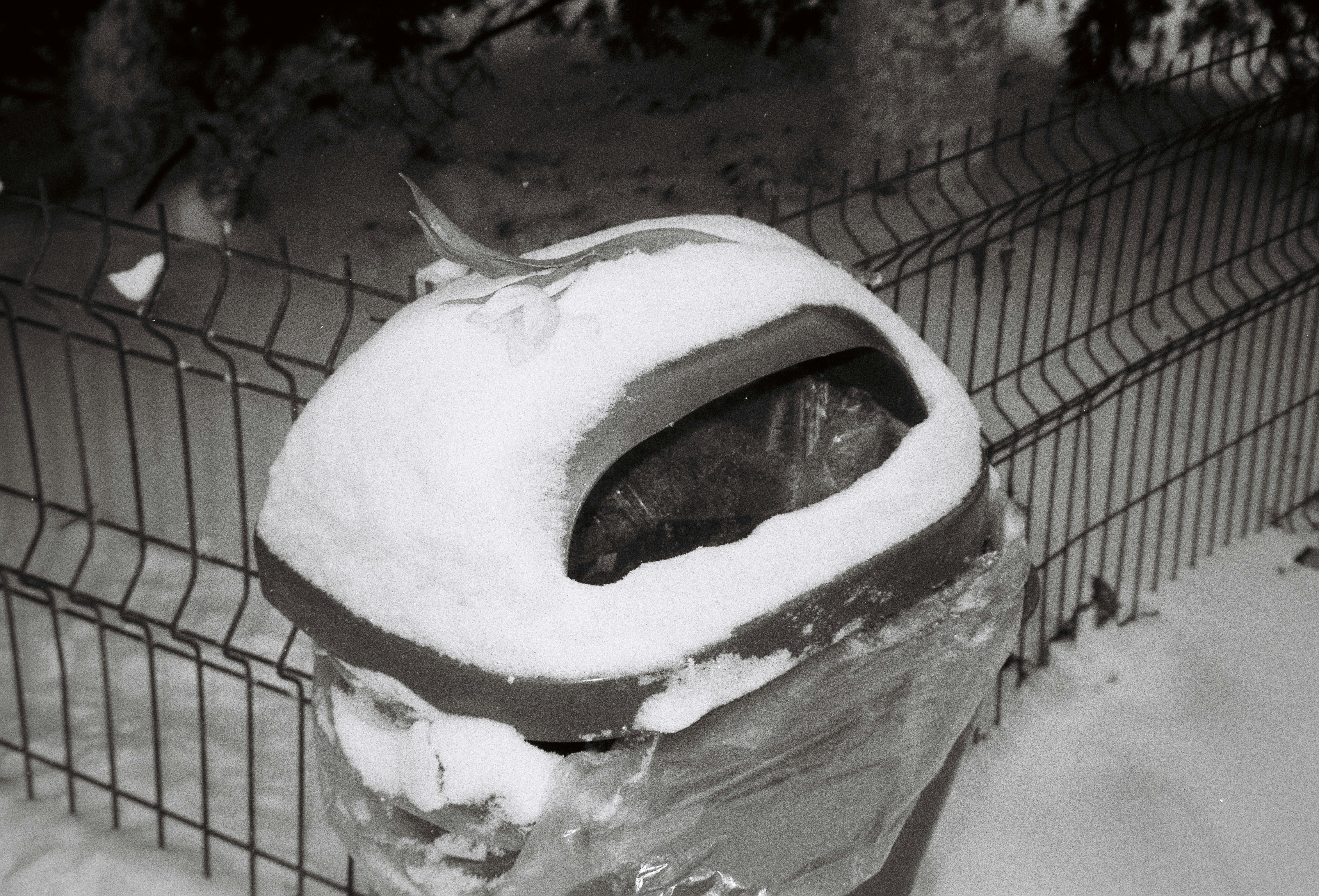 Snow covered trash can next to a fence.