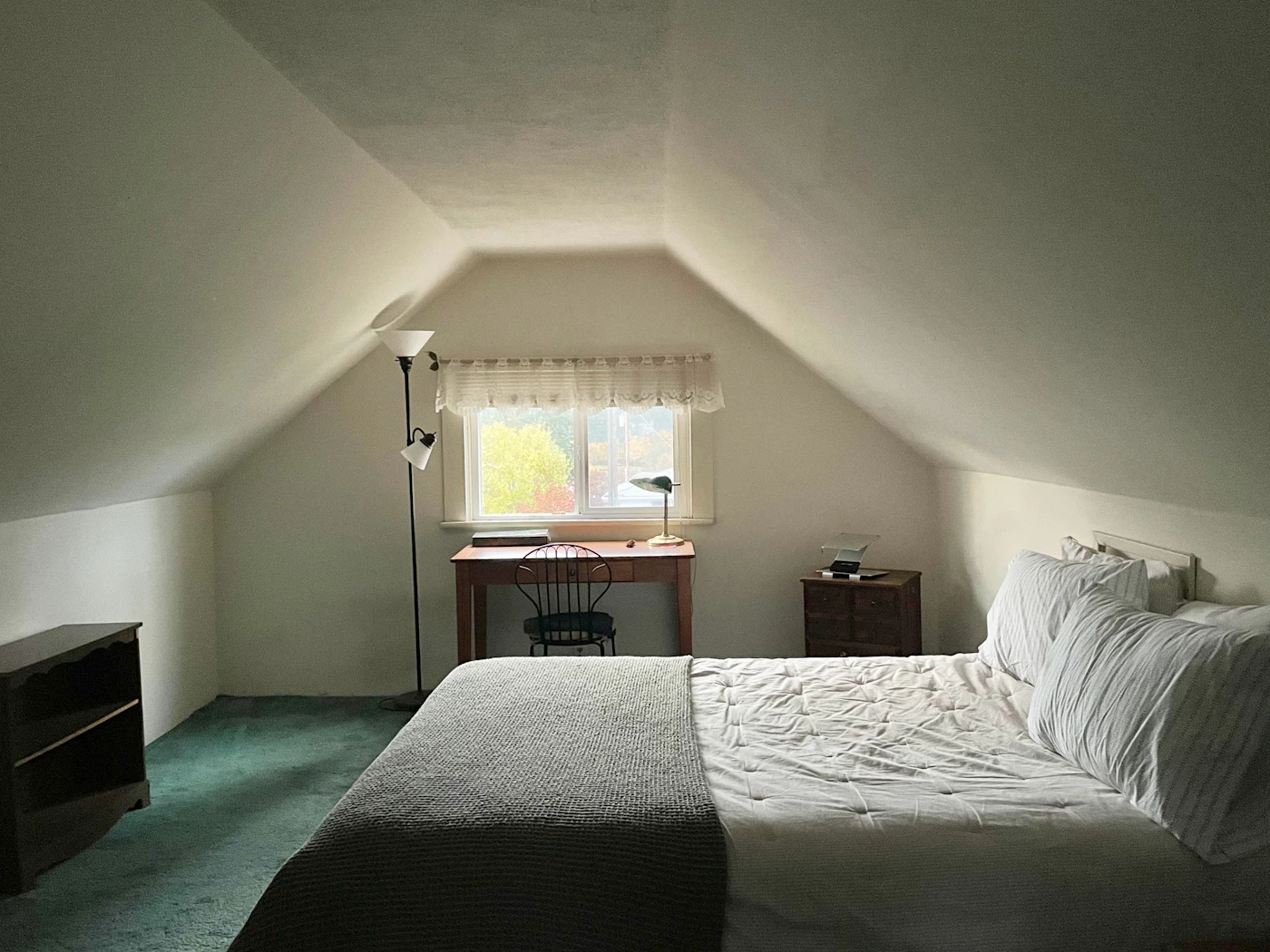 A simple dorm room with a single bed and a small wooden study desk and lamp set by the window