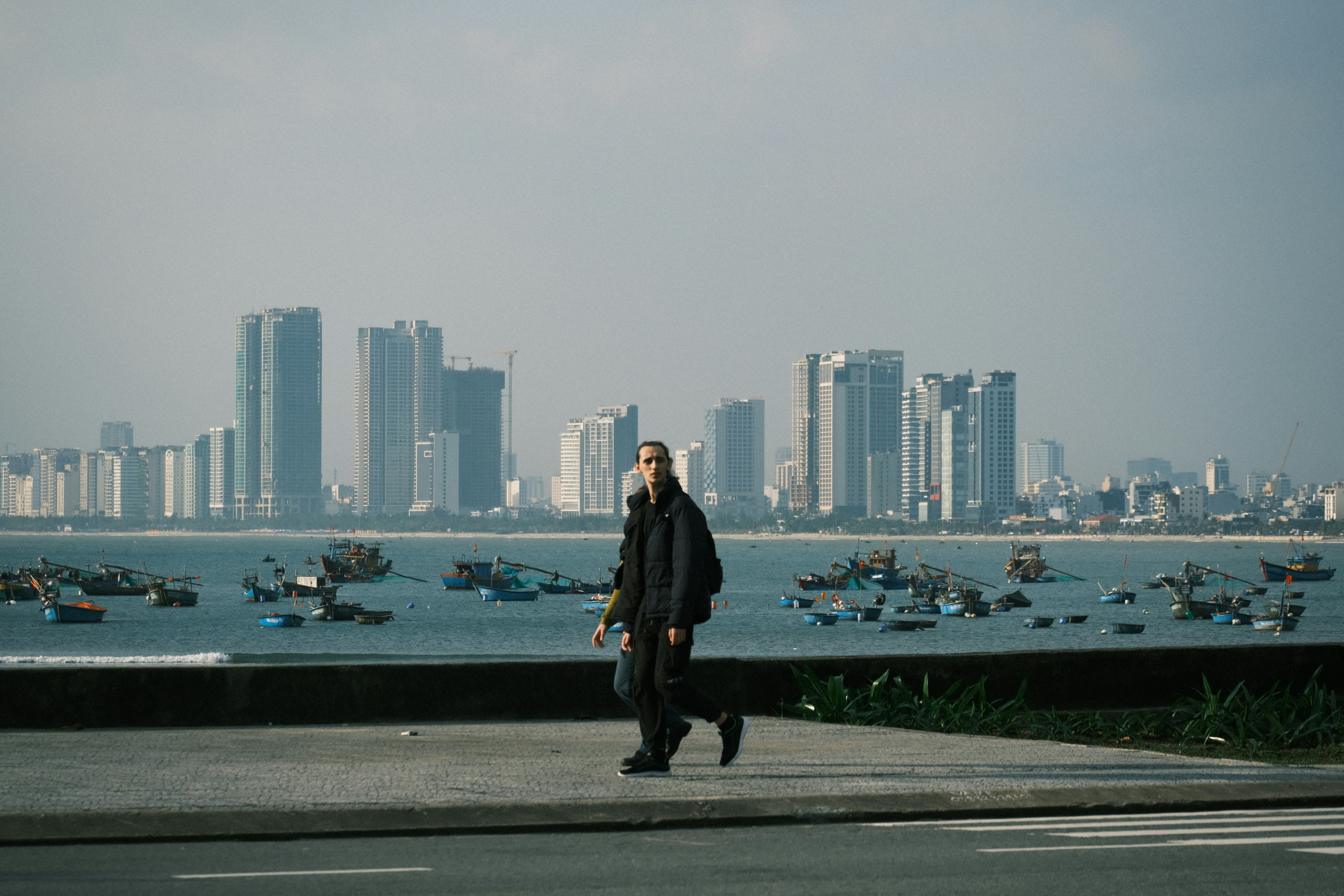 Person walks along waterfront with city skyline and boats.