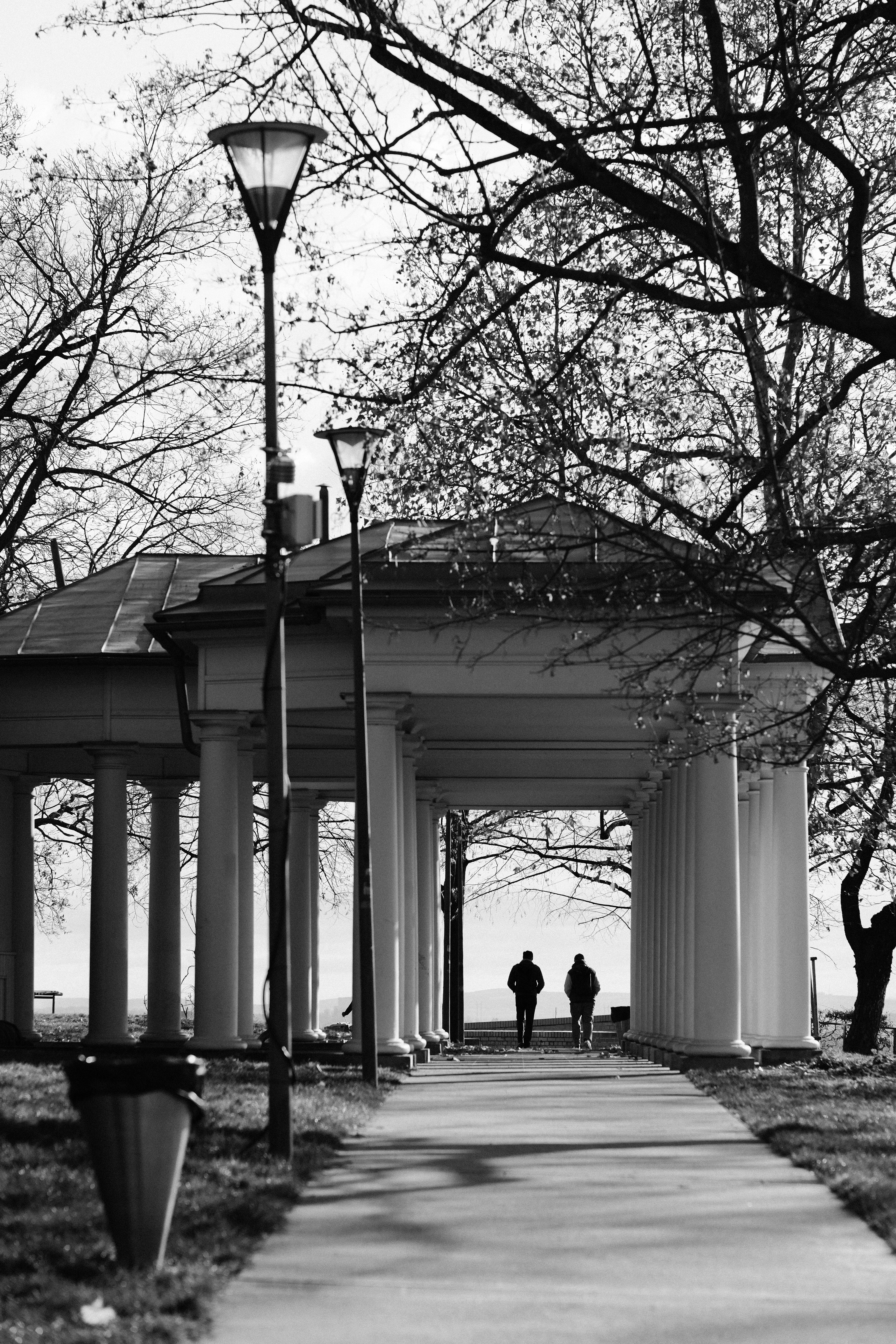 Two people walk through a colonnade by the water.