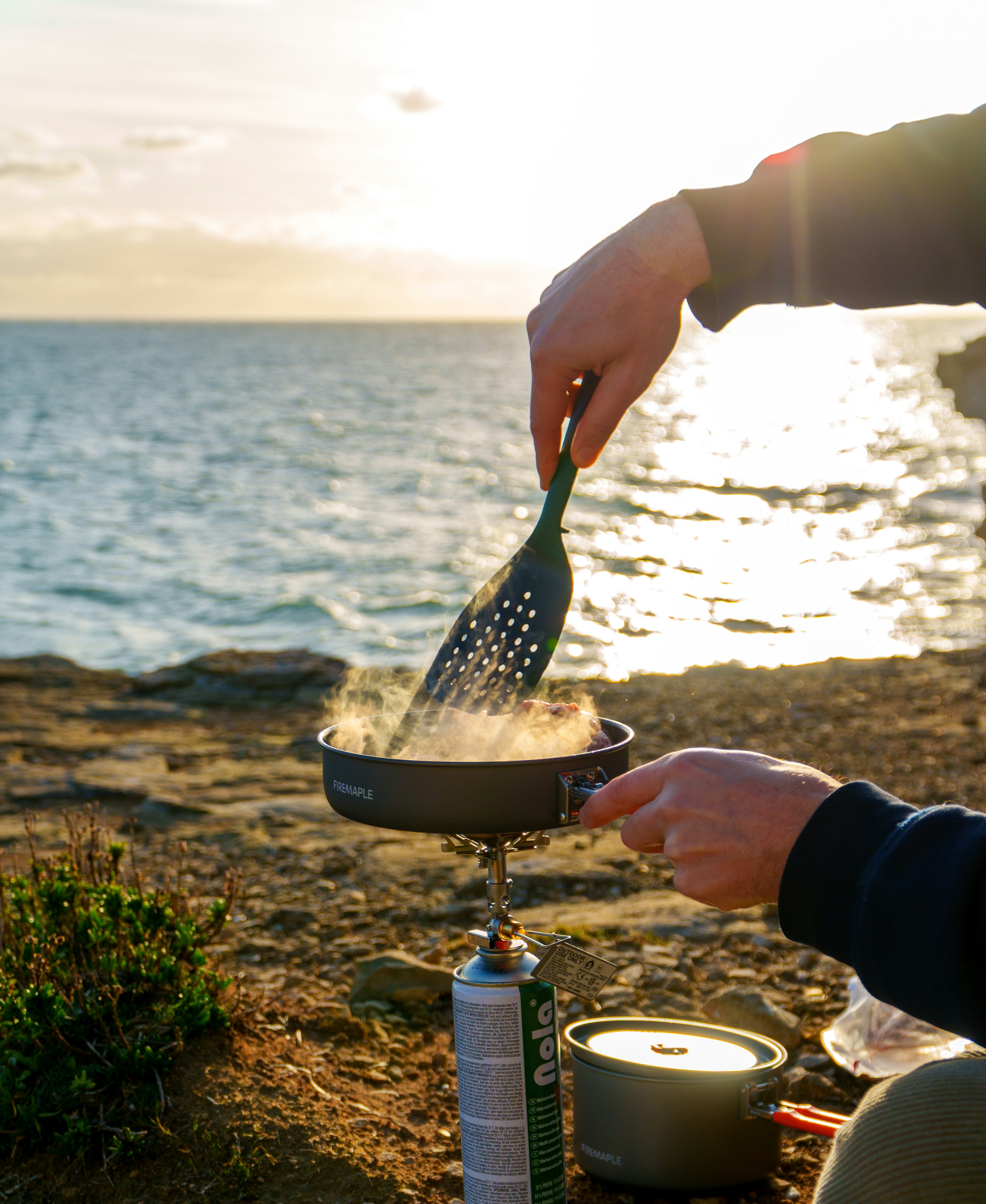 Person cooking outdoors by the ocean at sunset.