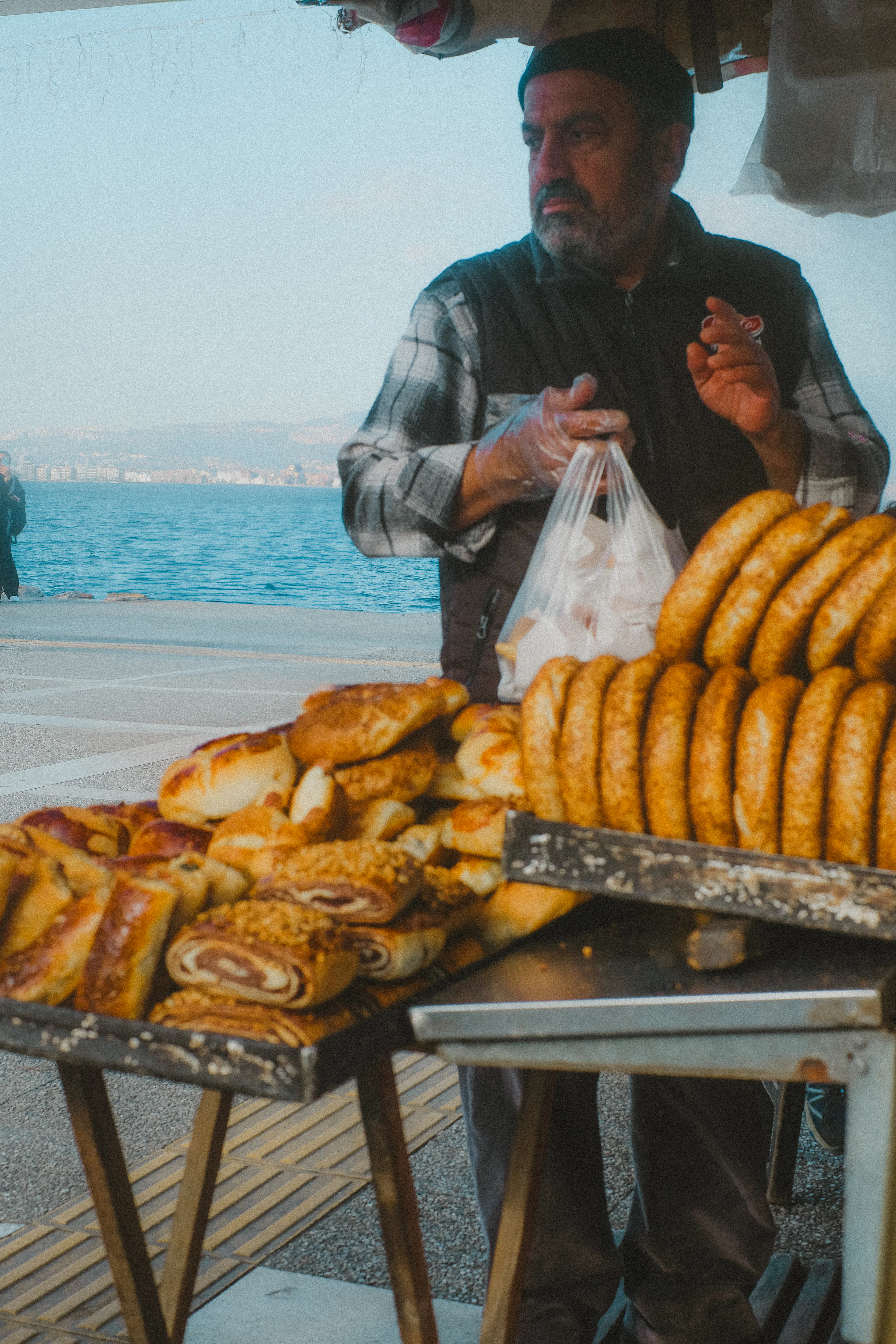 A street vendor in Izmir bagging pastries at his cart beside the water, sesame-crusted gevrek rings stacked in rows on a tray and flaky rolled boyoz and pogaca spread across the counter, the blue Gulf of Izmir and its far hills behind him