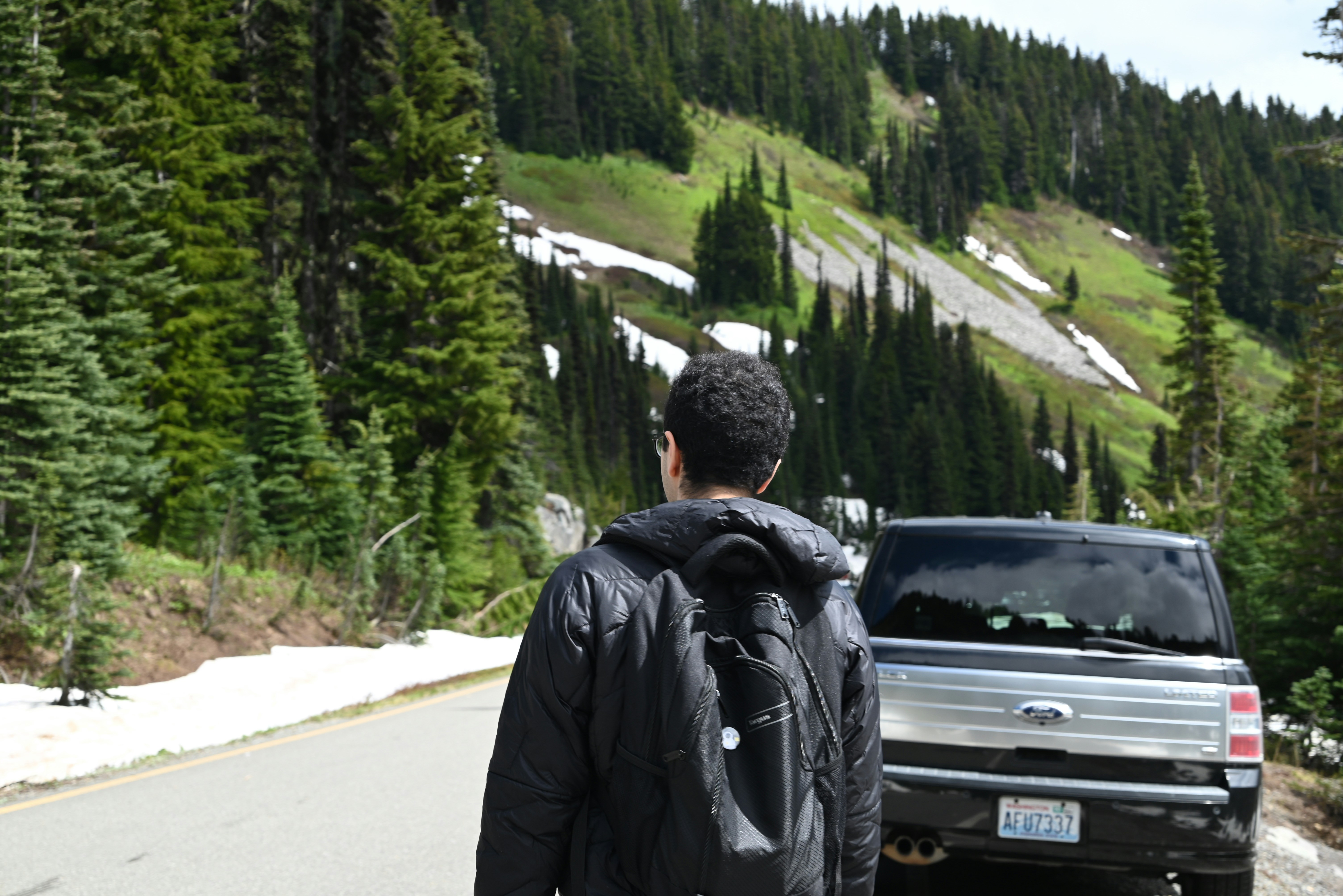 Man with backpack walks on road near forest and car.