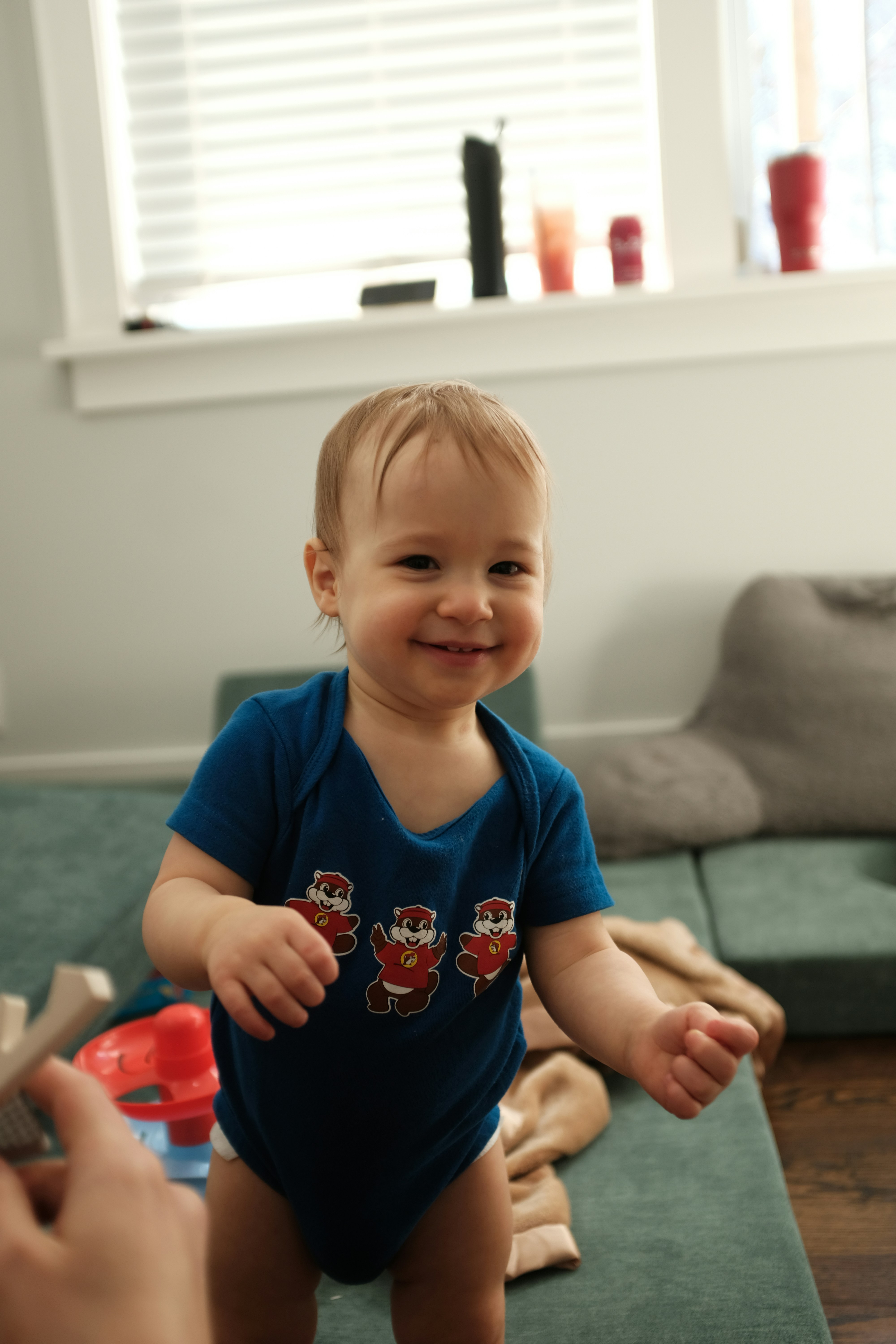 A smiling baby in a blue onesie stands indoors
