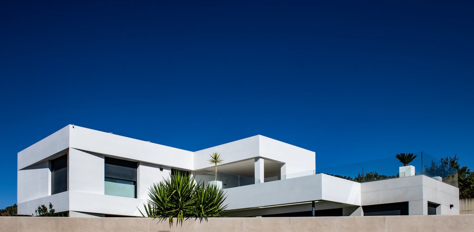 Modern white stucco building with flat roofline and parapet walls against a clear blue sky, showing roof-to-wall transition points