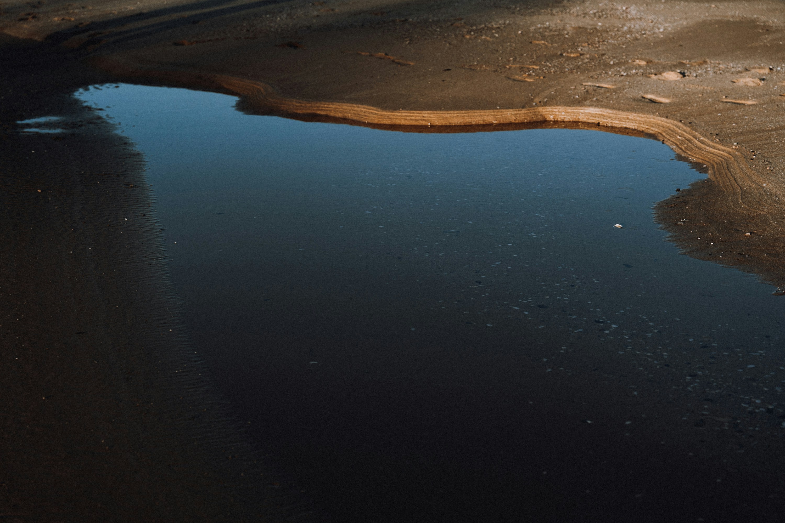A dark puddle reflects the sky on sandy ground.