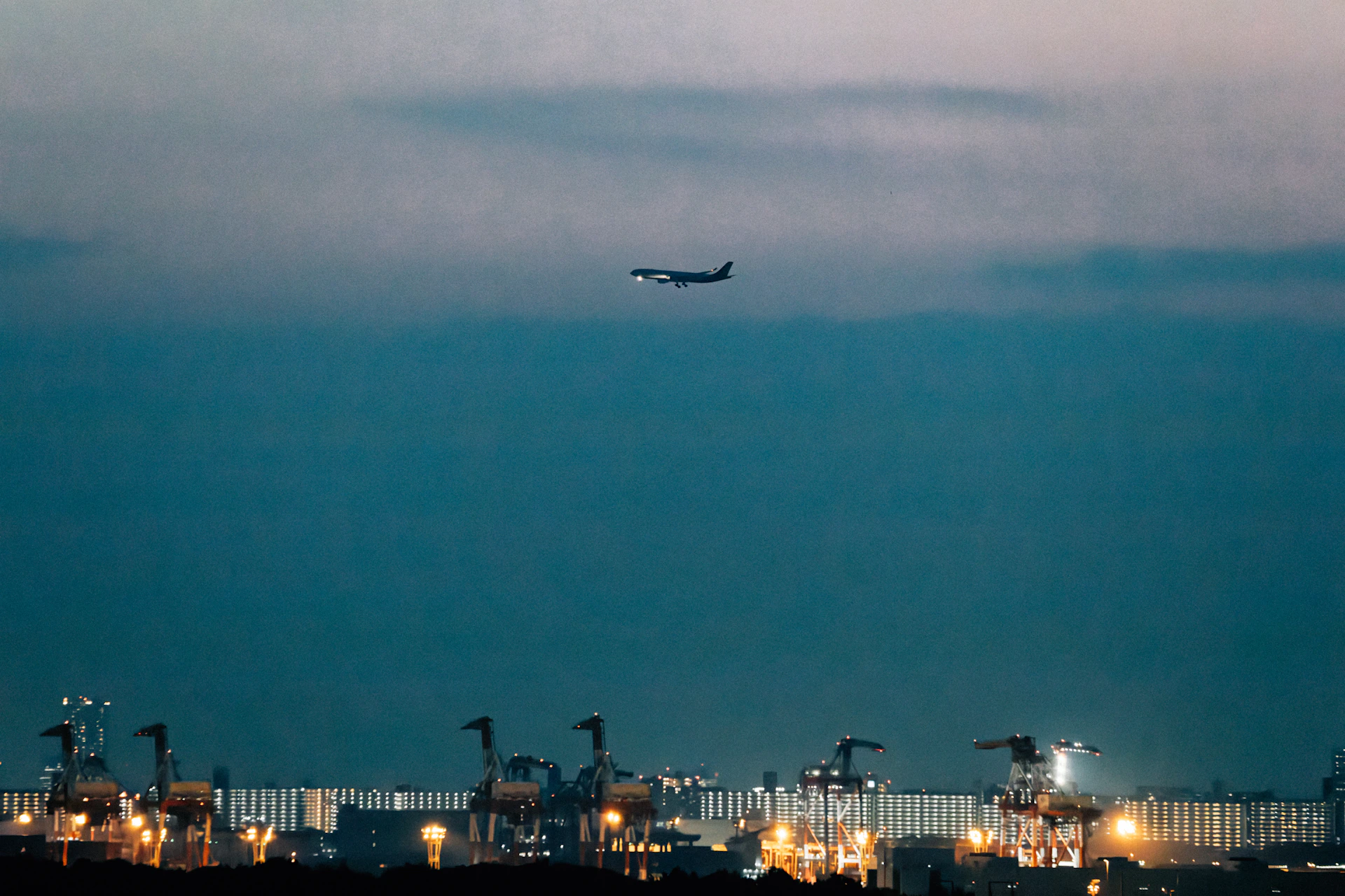 Airplane flying over illuminated industrial port at dusk.