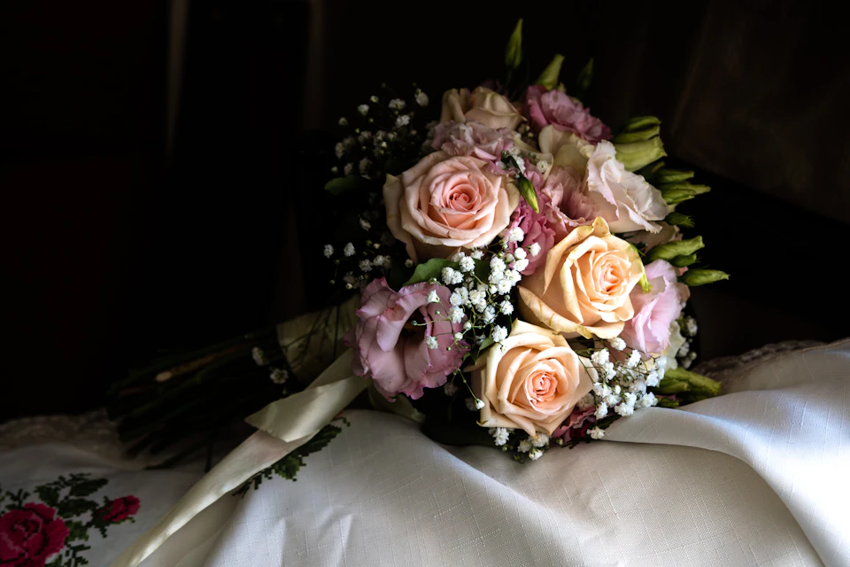 The image shows a bouquet of flowers resting on a white tablecloth, which is spread out and covering most of the frame. The bouquet consists of six roses with varying shades of pink and white petals, interspersed with baby's breath. The stems of the roses are visible through the gaps in the arrangement. The background appears to be a dark room or a dimly lit area, which contrasts with the vibrant colors of the flowers.