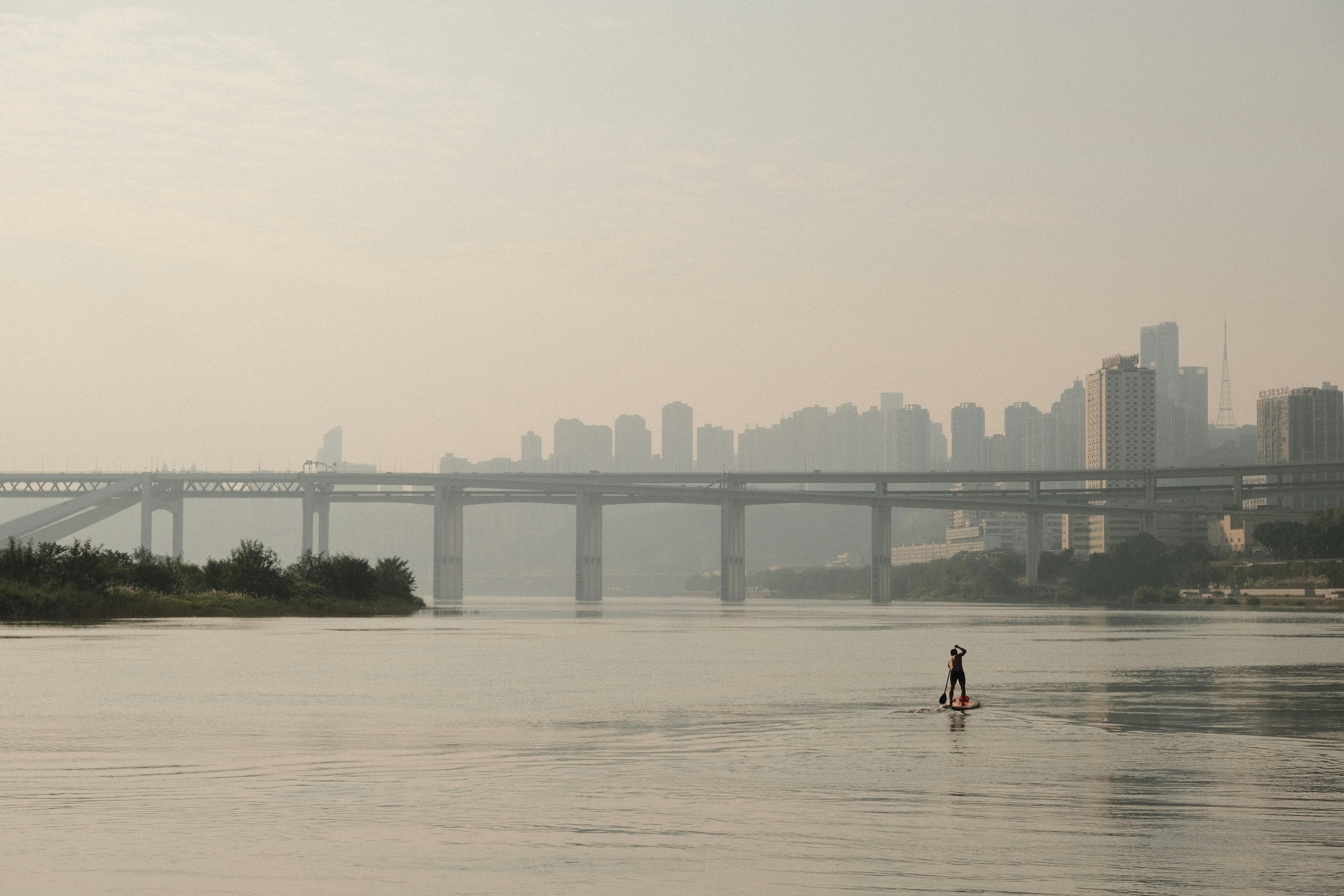 A lone paddleboarder on a wide river with city skyline.