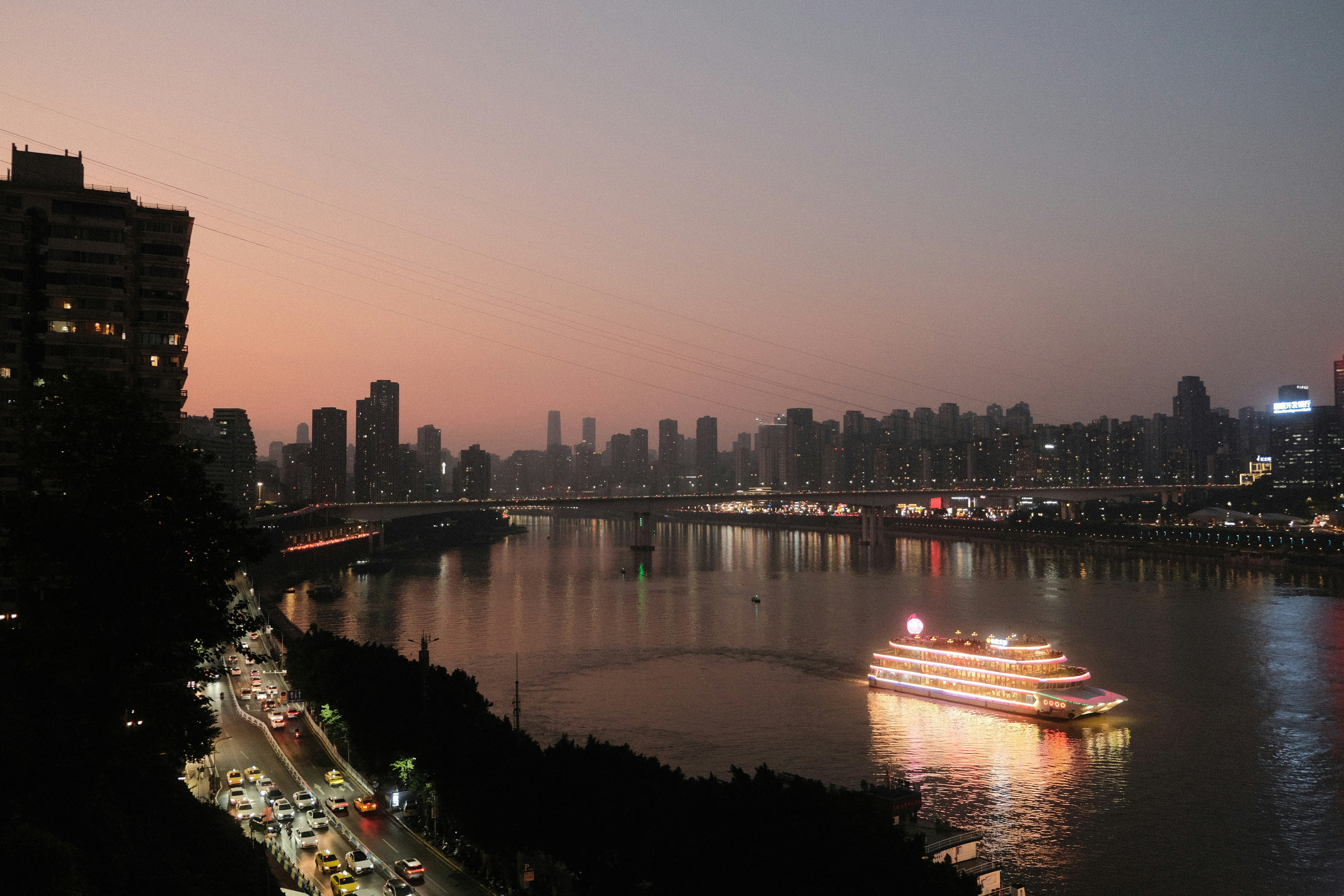 Illuminated boat sails on river past city skyline at dusk