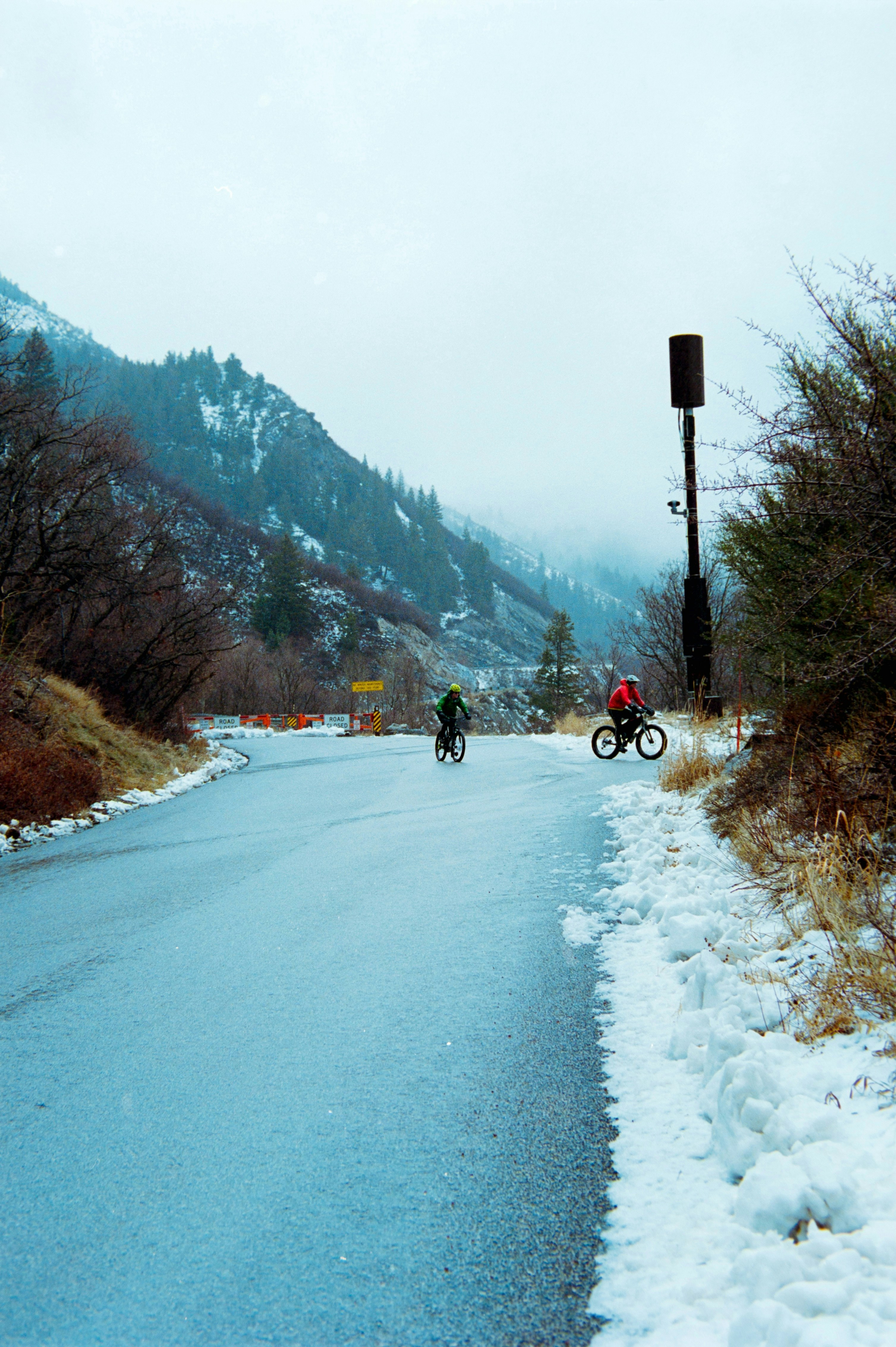 Two cyclists ride on a snowy road near mountains.