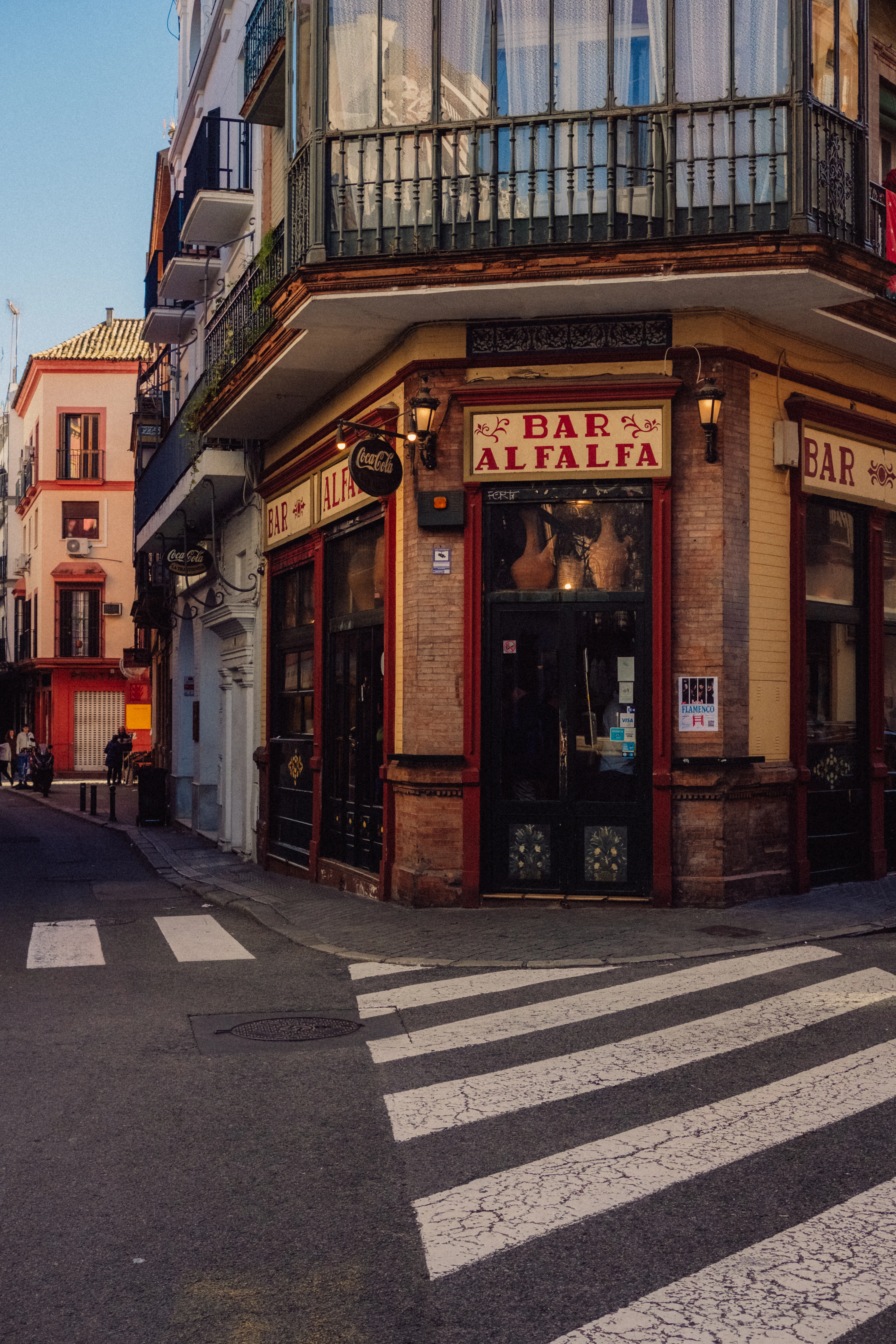 The corner facade of Bar Alfalfa, a small tapas bar in Seville, with wrought-iron balconies above and an empty crossing in front