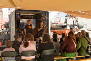 Two people present to an outdoor audience near boats.