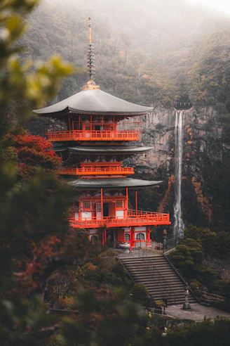 A vibrant red pagoda stands beside a tall waterfall.