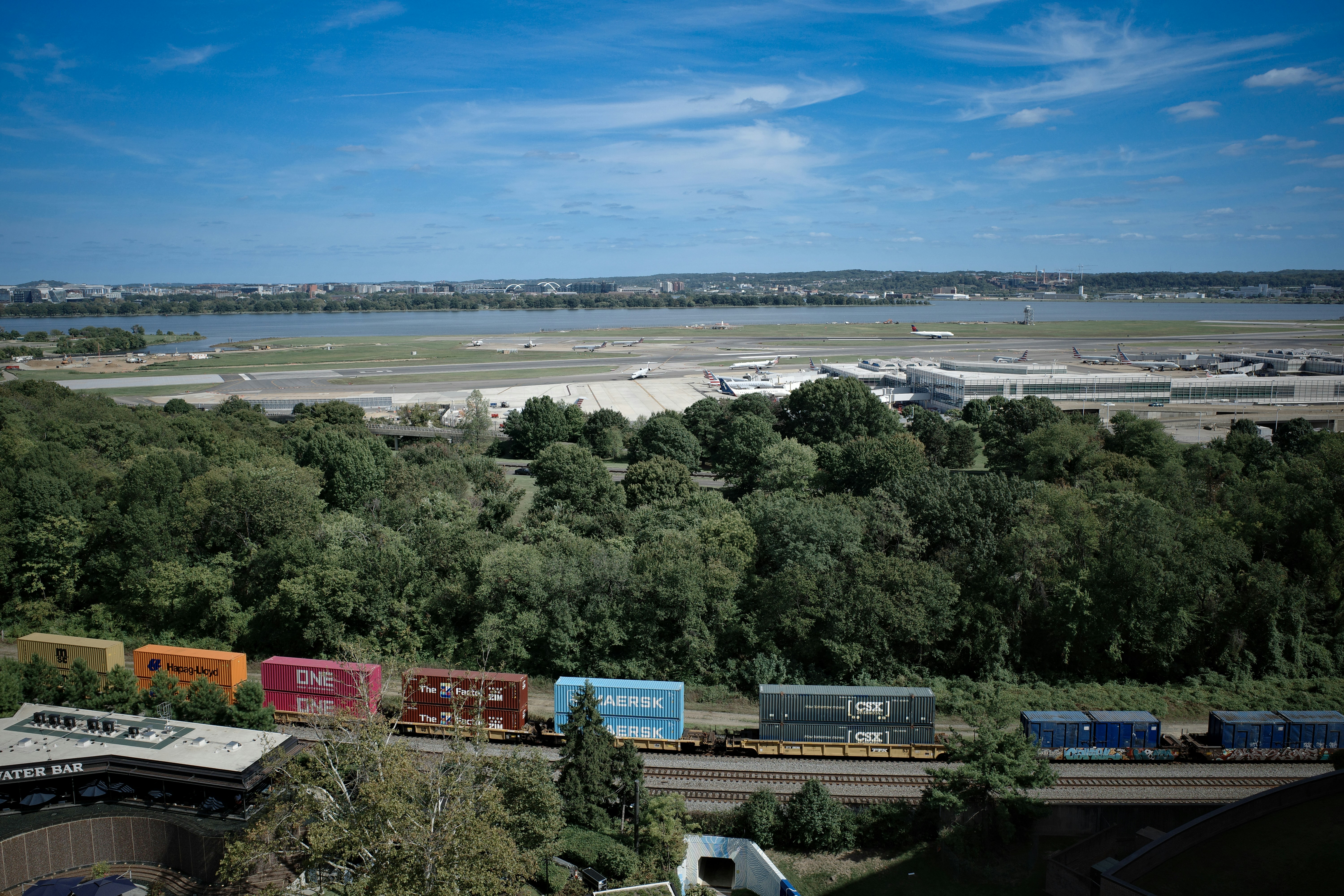 A freight train travels past trees towards a distant city.