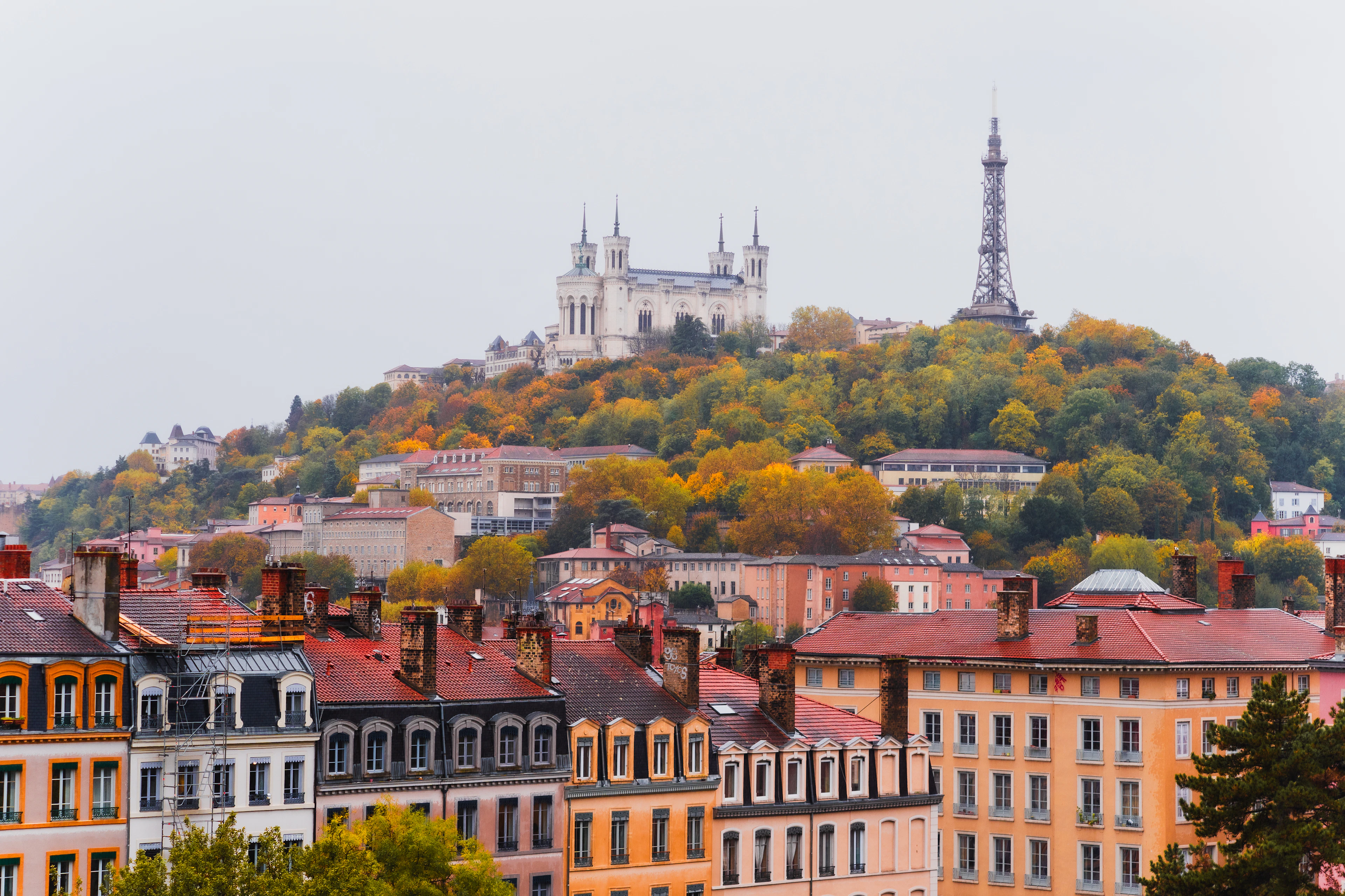 The white towers of the Fourviere basilica and the iron Fourviere tower rising above an autumn-coloured hillside in Lyon, with a row of ochre and pink apartment facades and red roofs along the foot of the slope