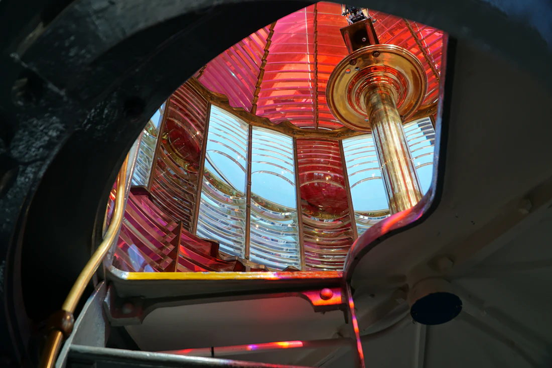 Looking up through the hatch of a lighthouse lamp room into a Fresnel lens of red and clear glass rings around a brass column
