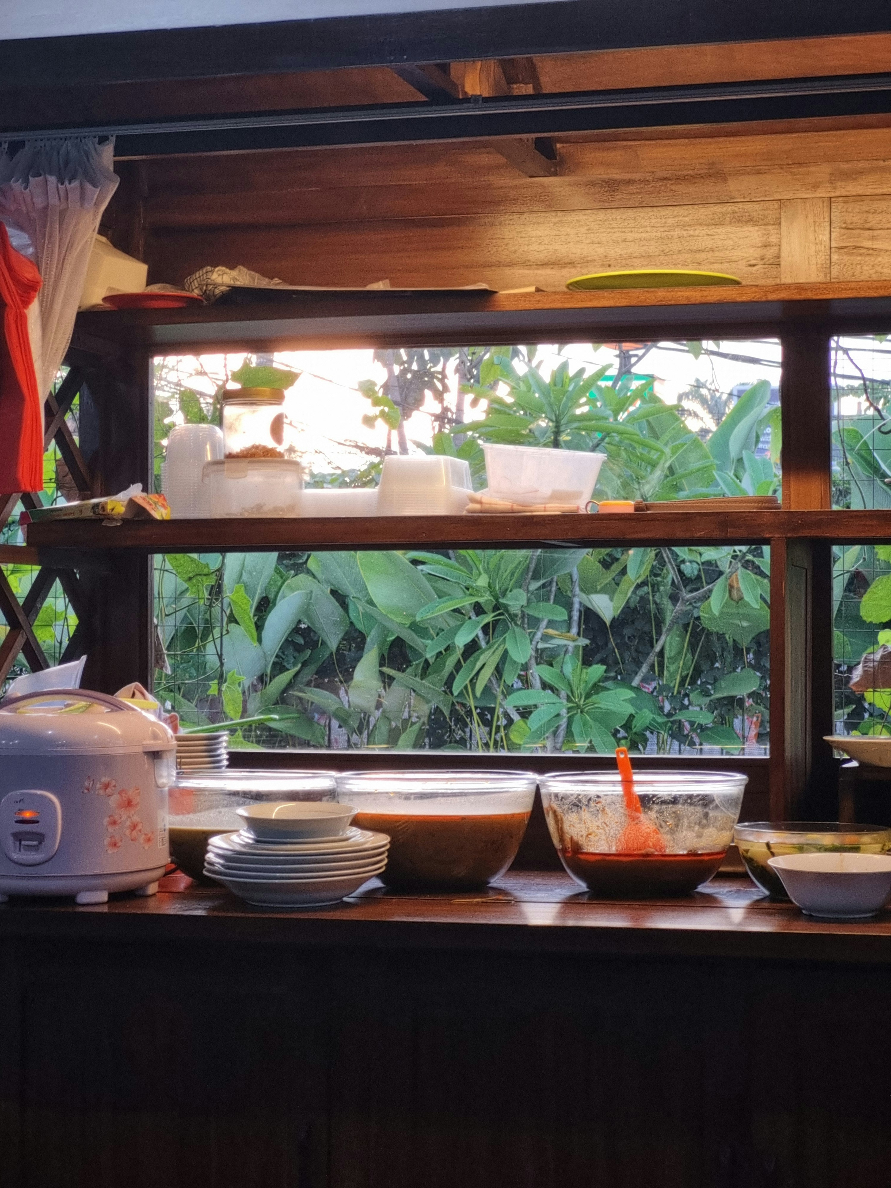 A warm and inviting scene in a semi-outdoor dining area. Glass bowls filled with savory dishes and a rice cooker sit ready on a wooden counter, framed by a large window revealing lush tropical greenery outside. A peaceful culinary moment capturing the essence of authentic home-style dining and slow living.