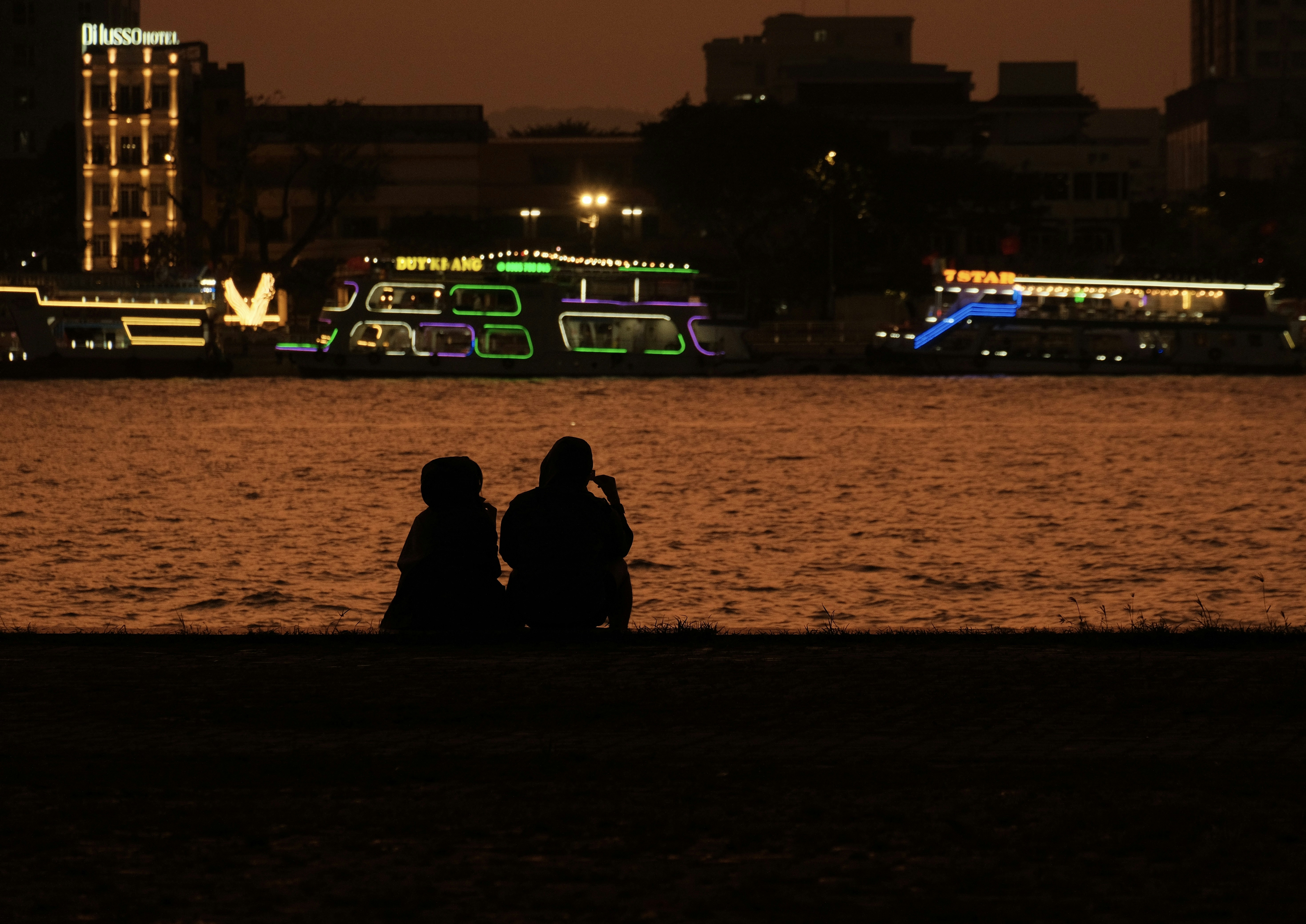 Two people watch illuminated boats on a river at night.