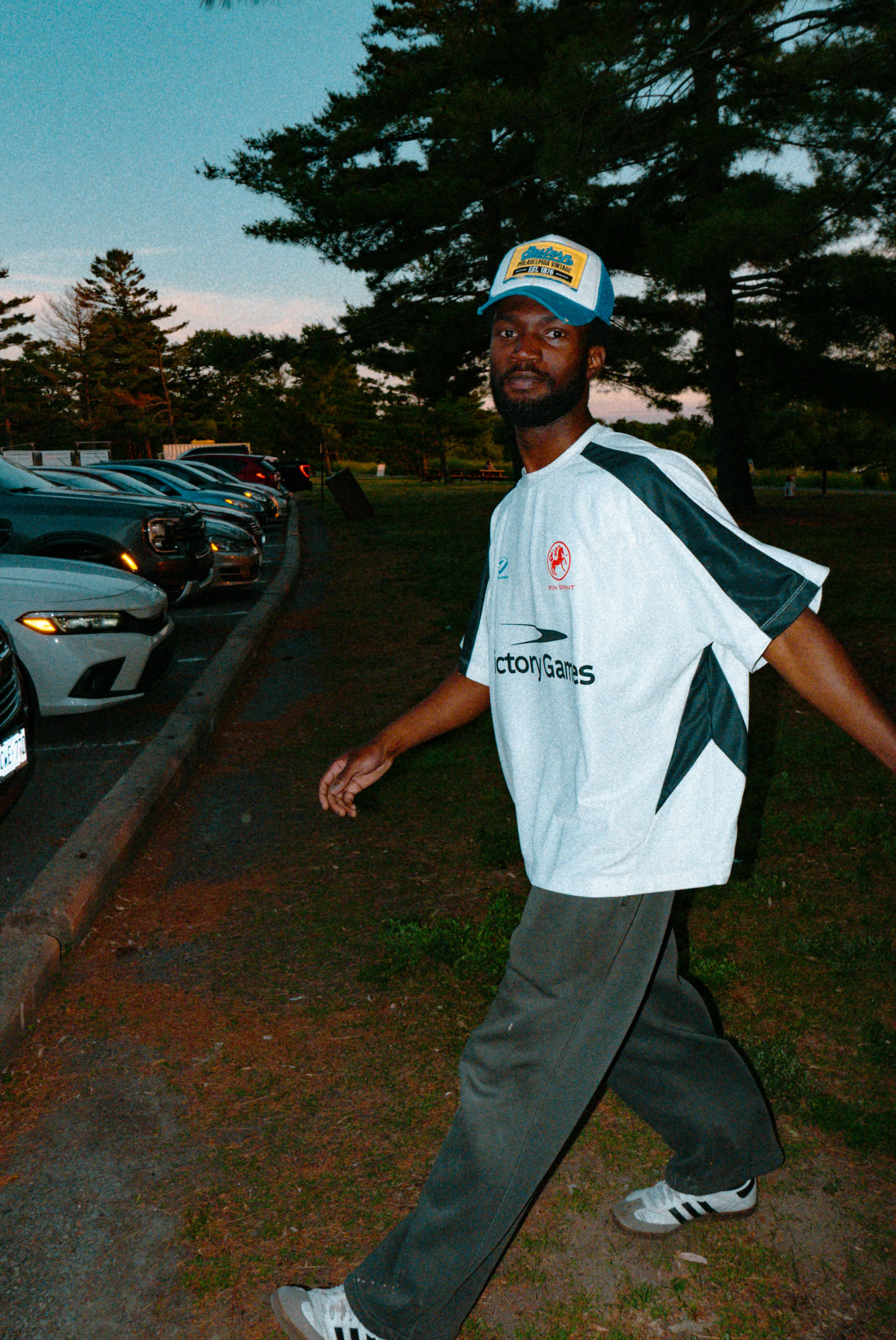 A man in a jersey and hat walks near cars.