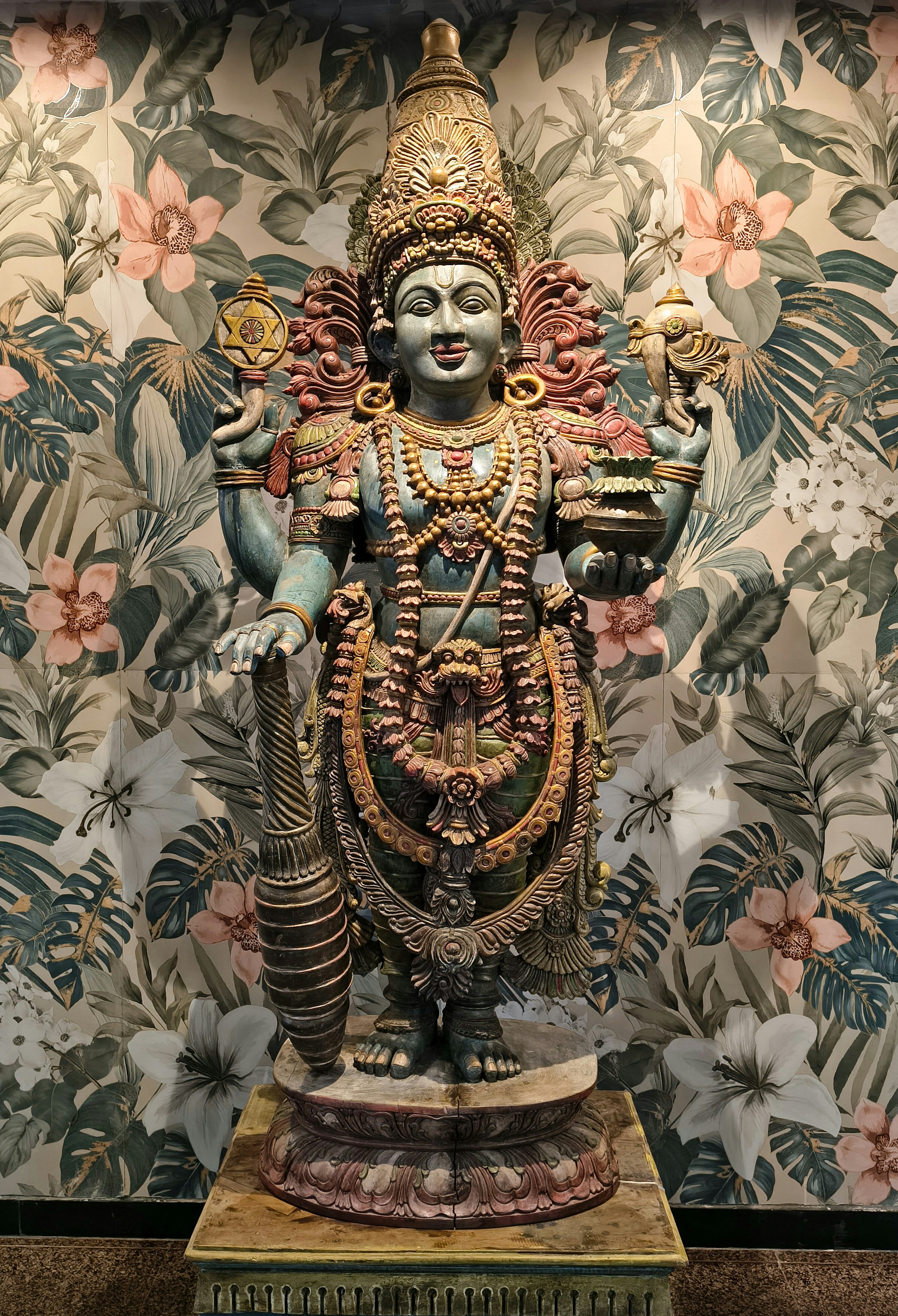 Ornate statue of a multi-armed deity against floral wallpaper.
