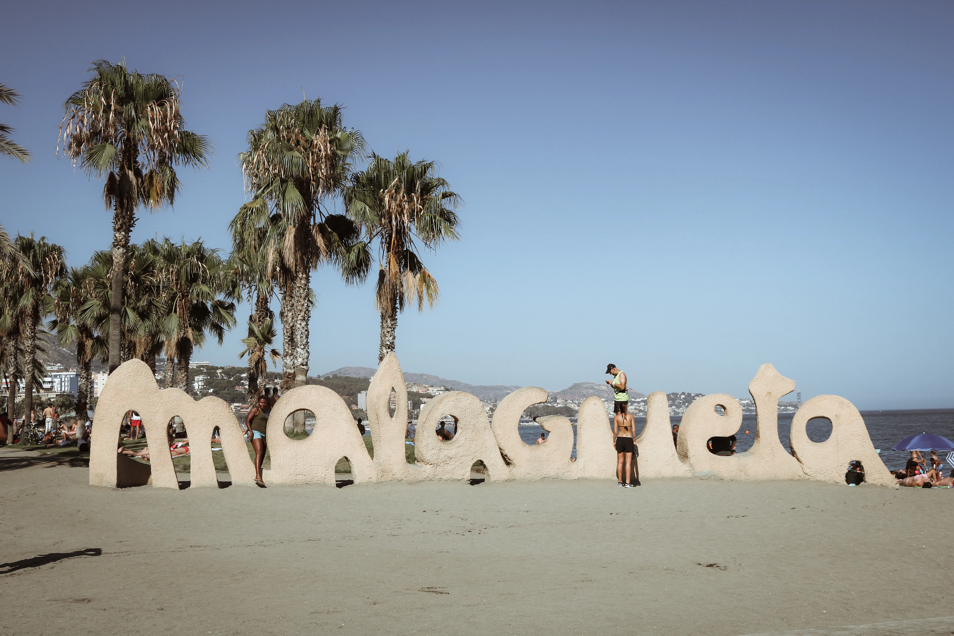 The giant sand-coloured malagueta letters standing on La Malagueta beach in Malaga, backed by a row of tall palms and the sea, with sunbathers scattered on the sand on a cloudless afternoon