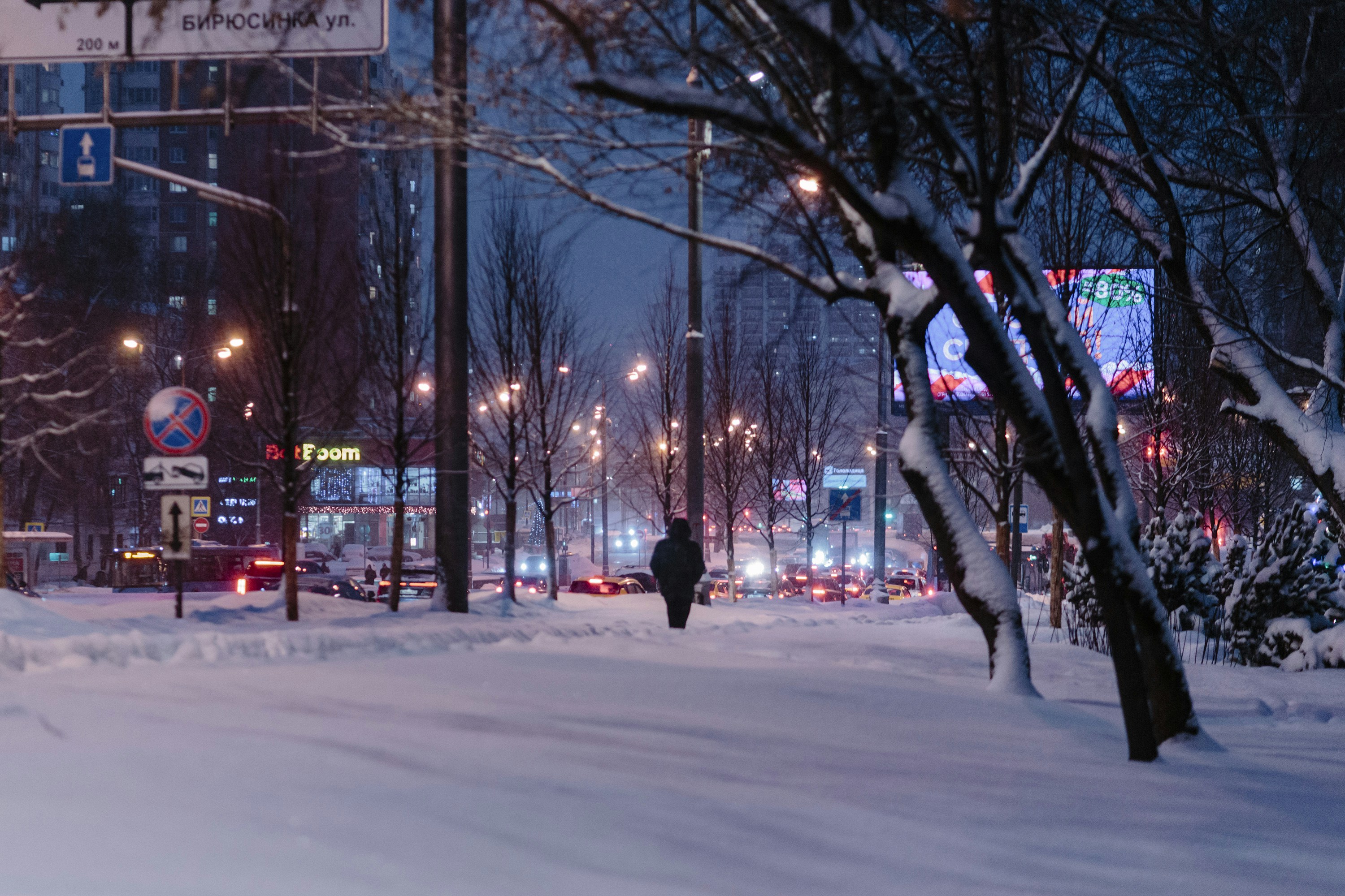 A lone figure walks down a snow-covered street at night.