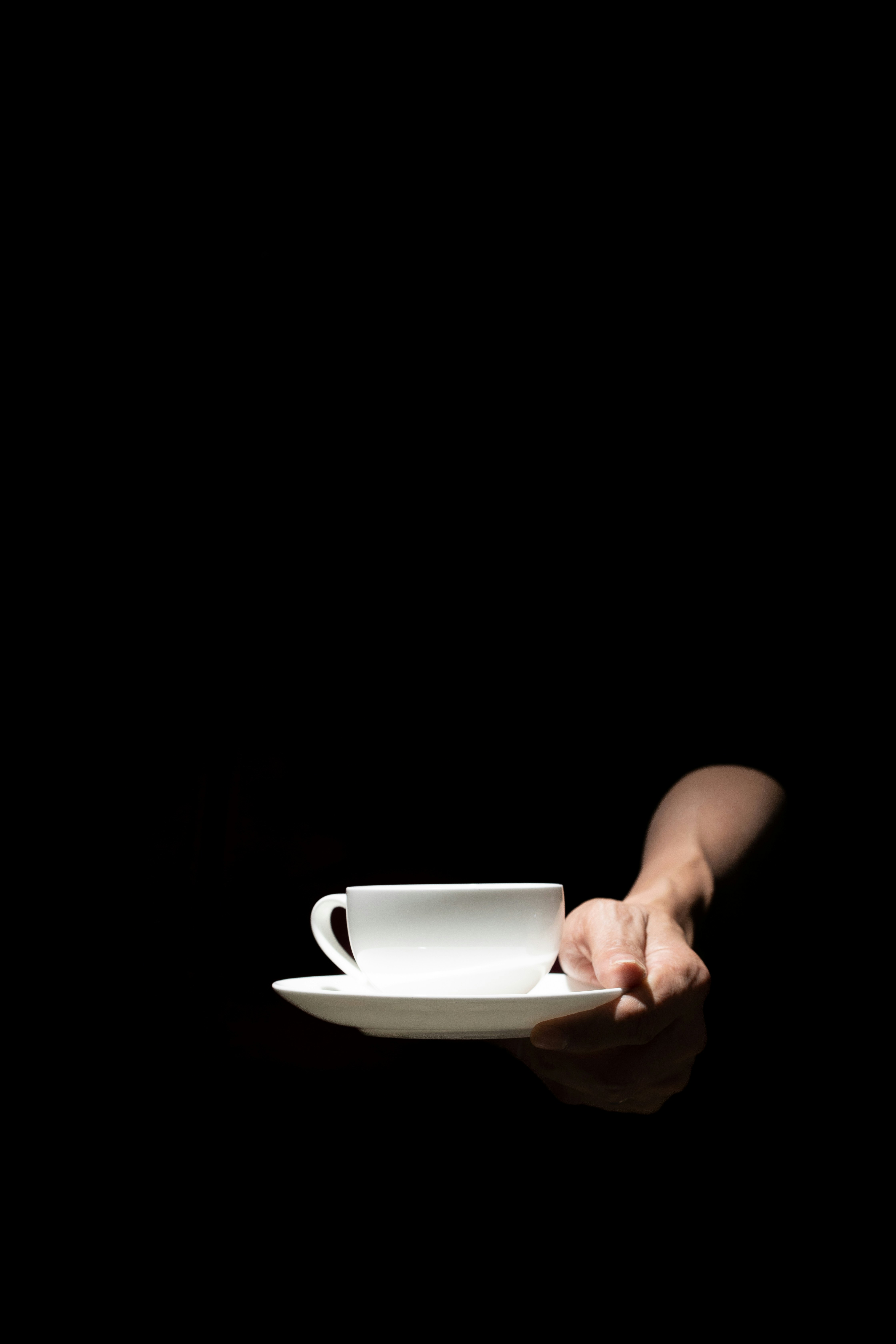 A hand holds a white teacup and saucer against black.
