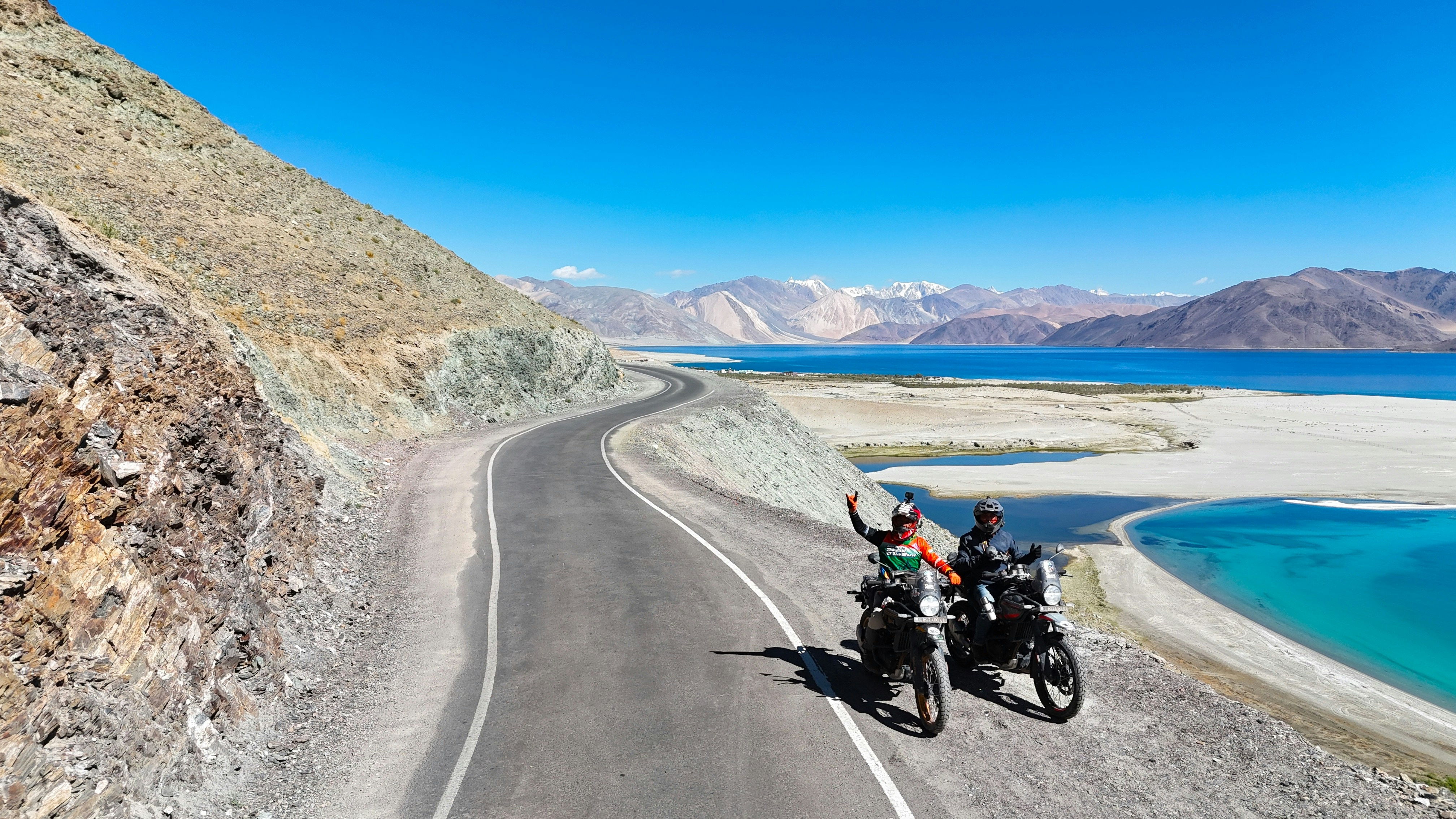 Two motorcyclists wave on a winding road by a lake.