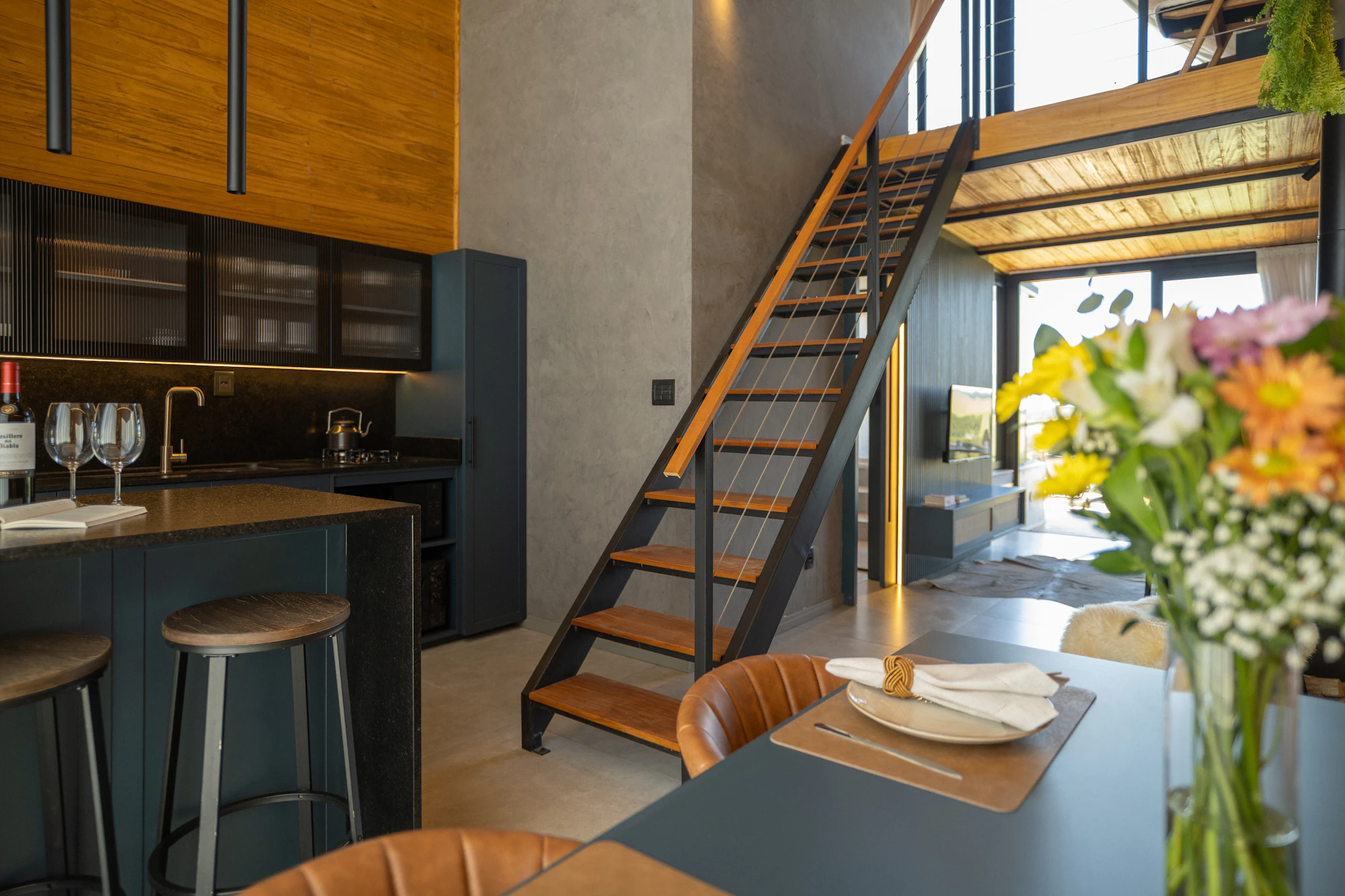 A modern open-plan kitchen and dining area features dark cabinetry, wood accents, a stone island, bar stools, and a staircase.