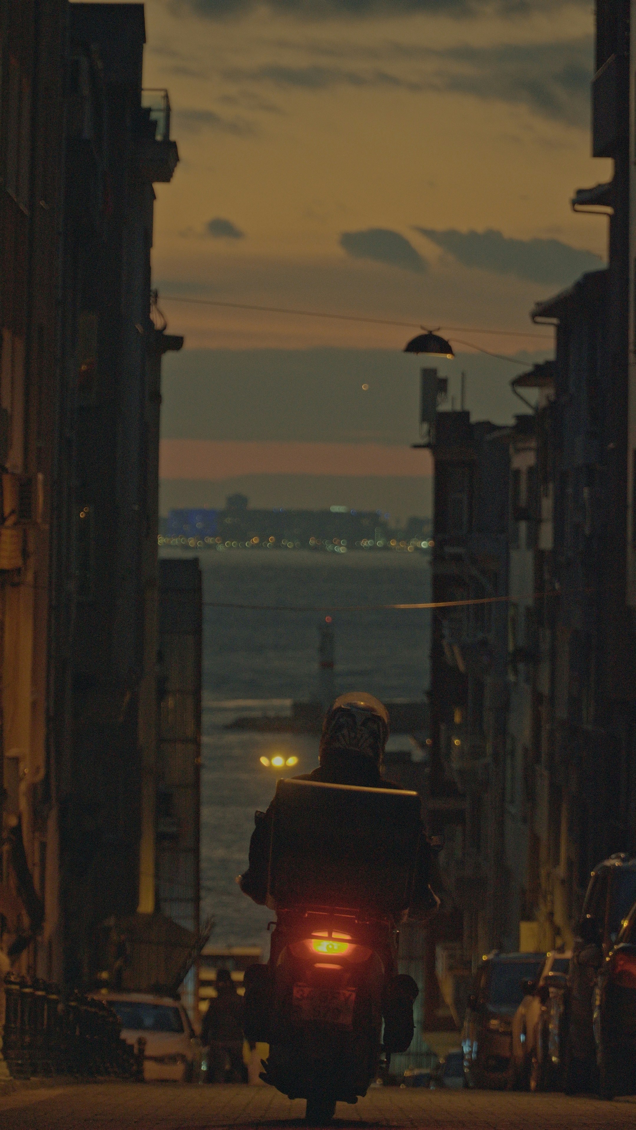 Motorcyclist with delivery box rides down street at dusk.