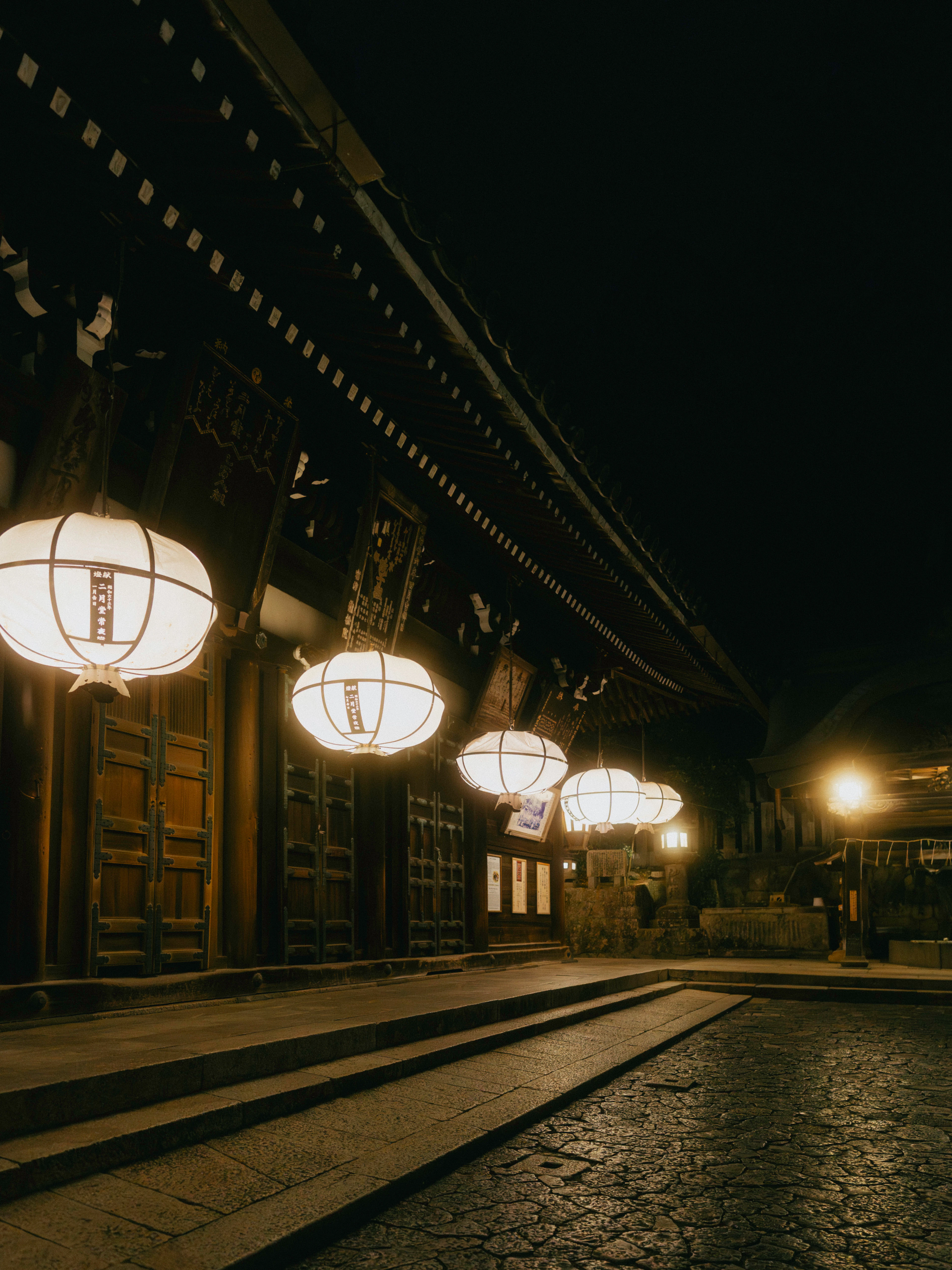 Large white paper lanterns burning under the eaves of Nigatsu-do at Todai-ji in Nara late at night, their light falling across dark timber doors and the worn stone steps below.