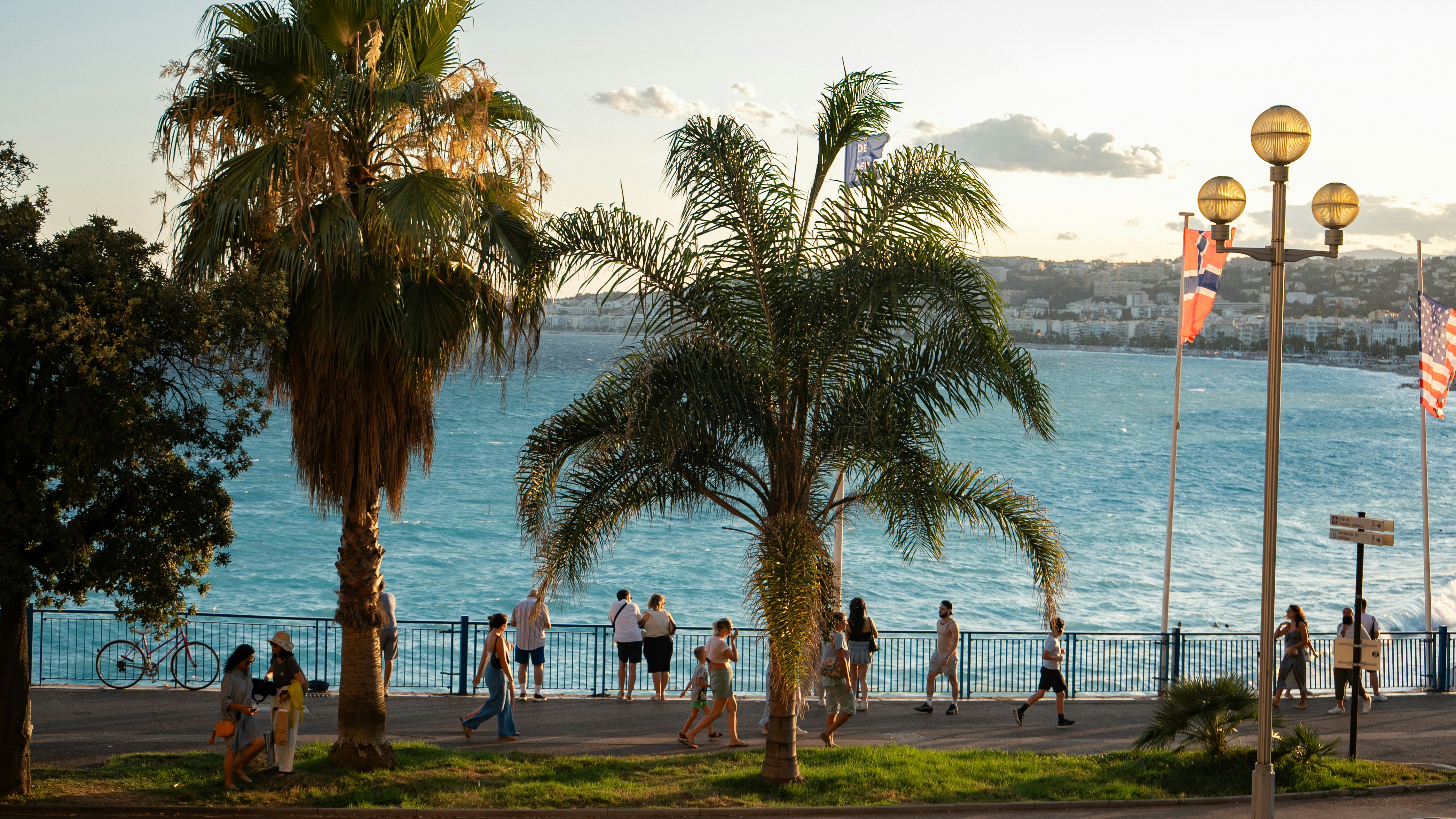 People stroll along a palm-lined promenade by the sea.