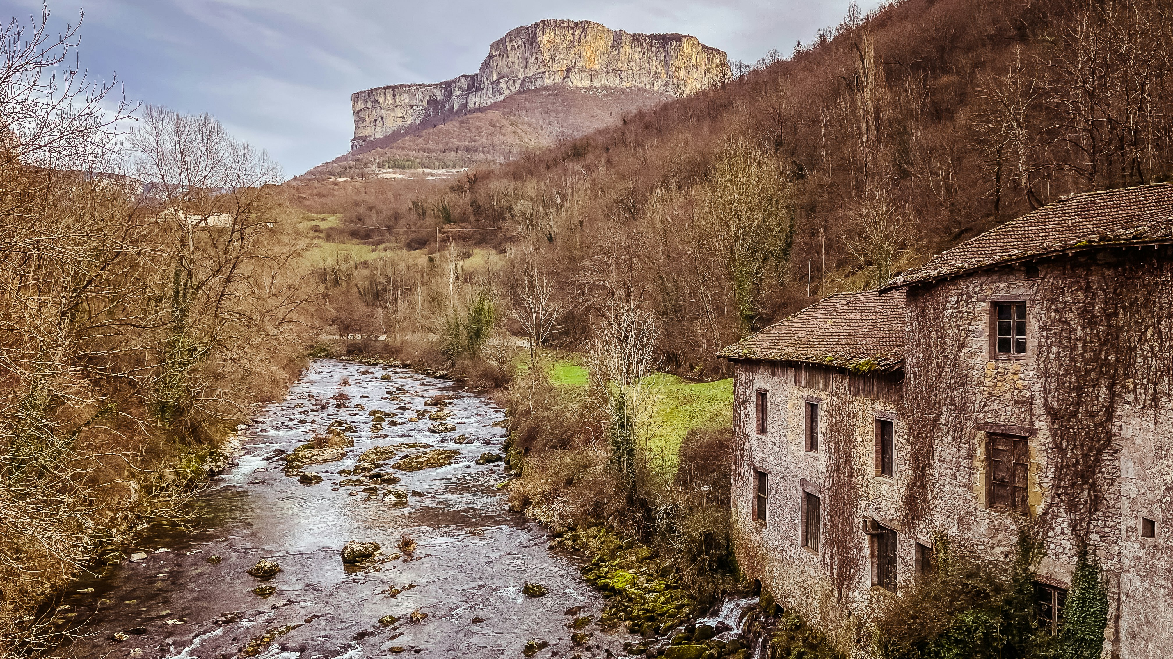 River flows past an old stone building near mountains.