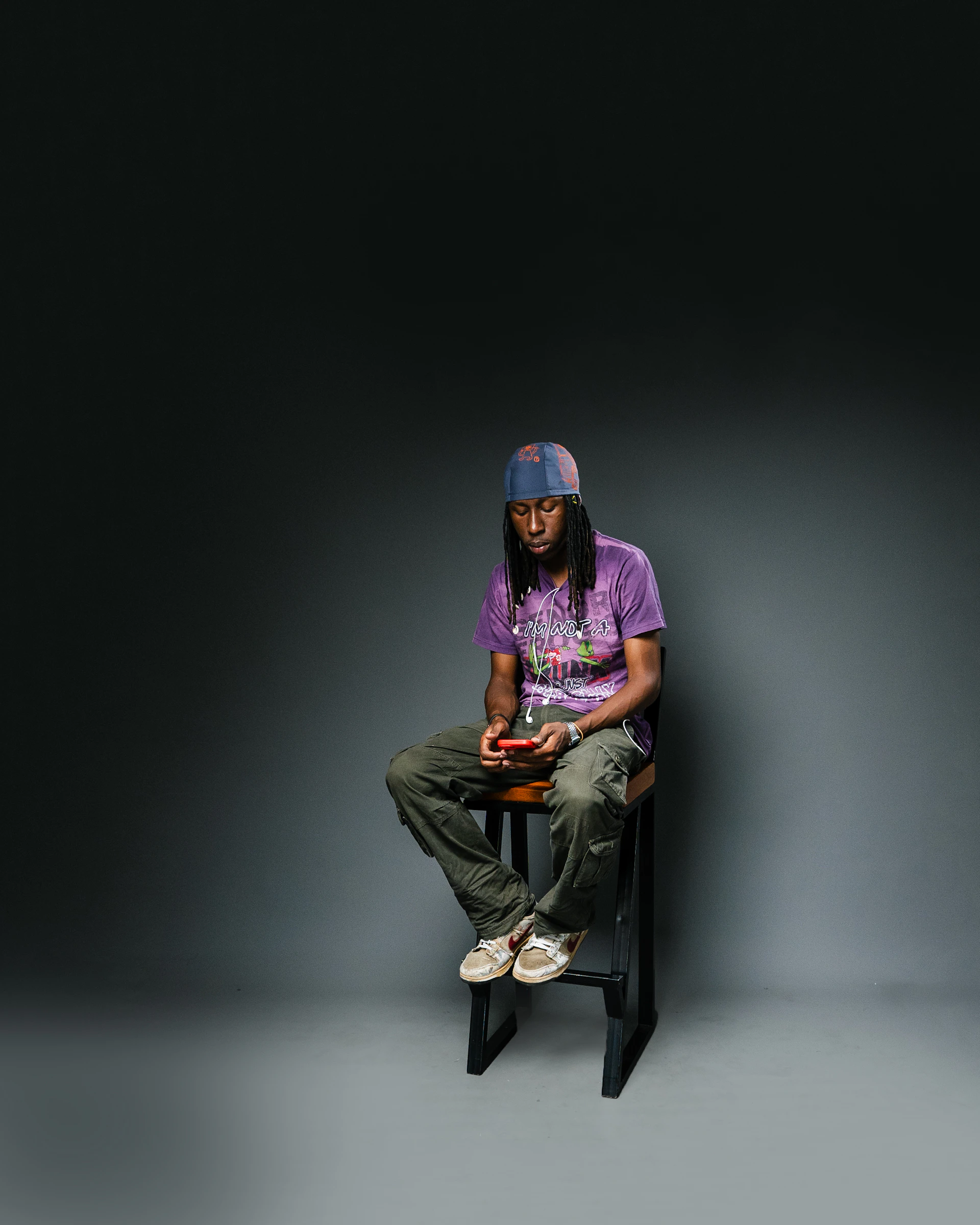 Man with dreadlocks wearing a hat and purple shirt sits.