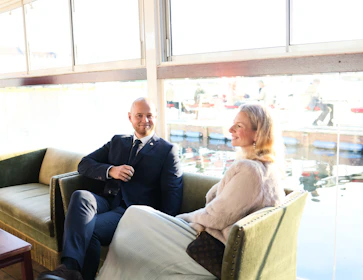 A man and woman sitting on a couch by window.